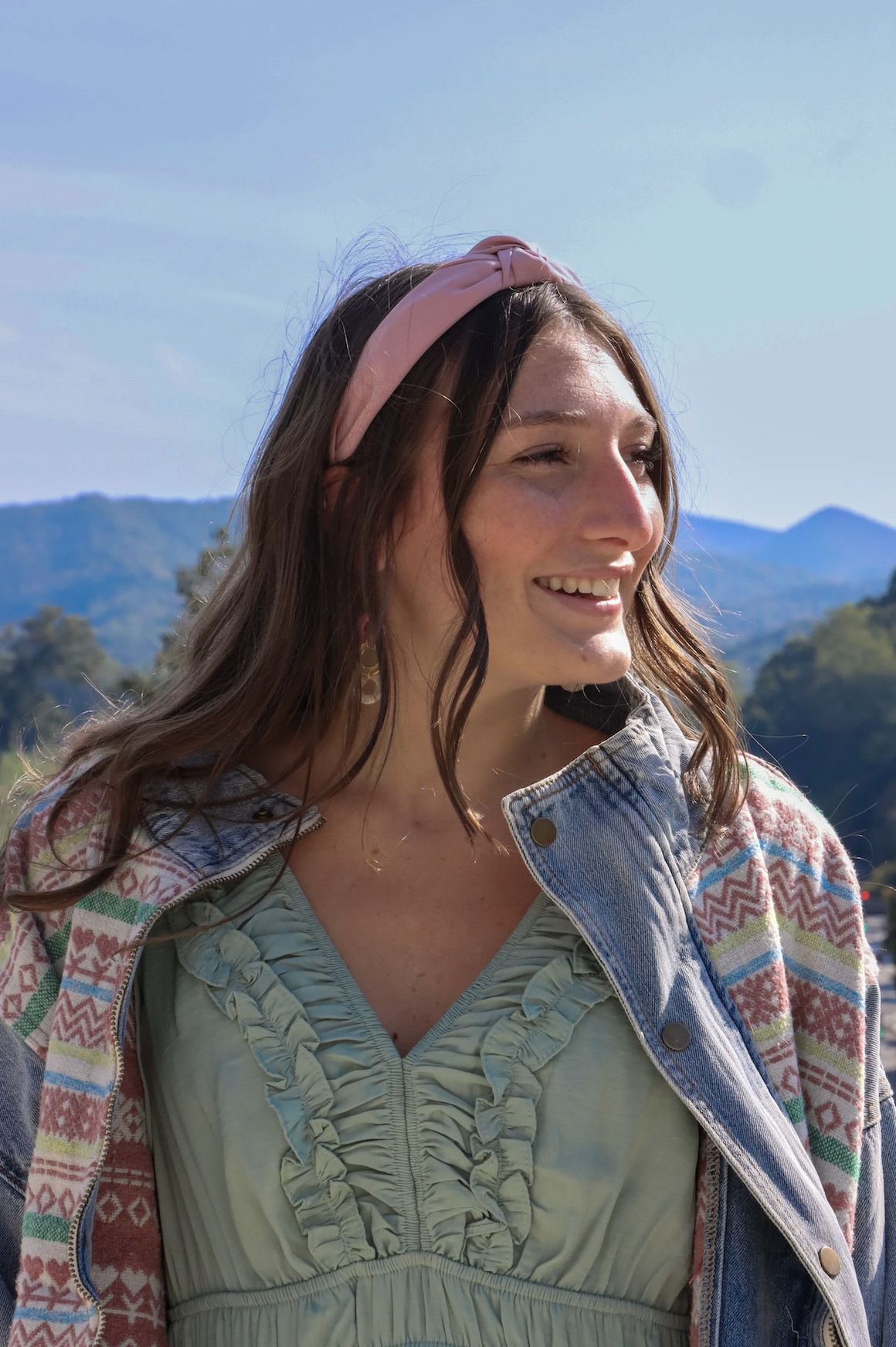 Young woman smiling outdoors with mountains in the background, wearing a pink headband, patterned jacket, and ruffled green top.