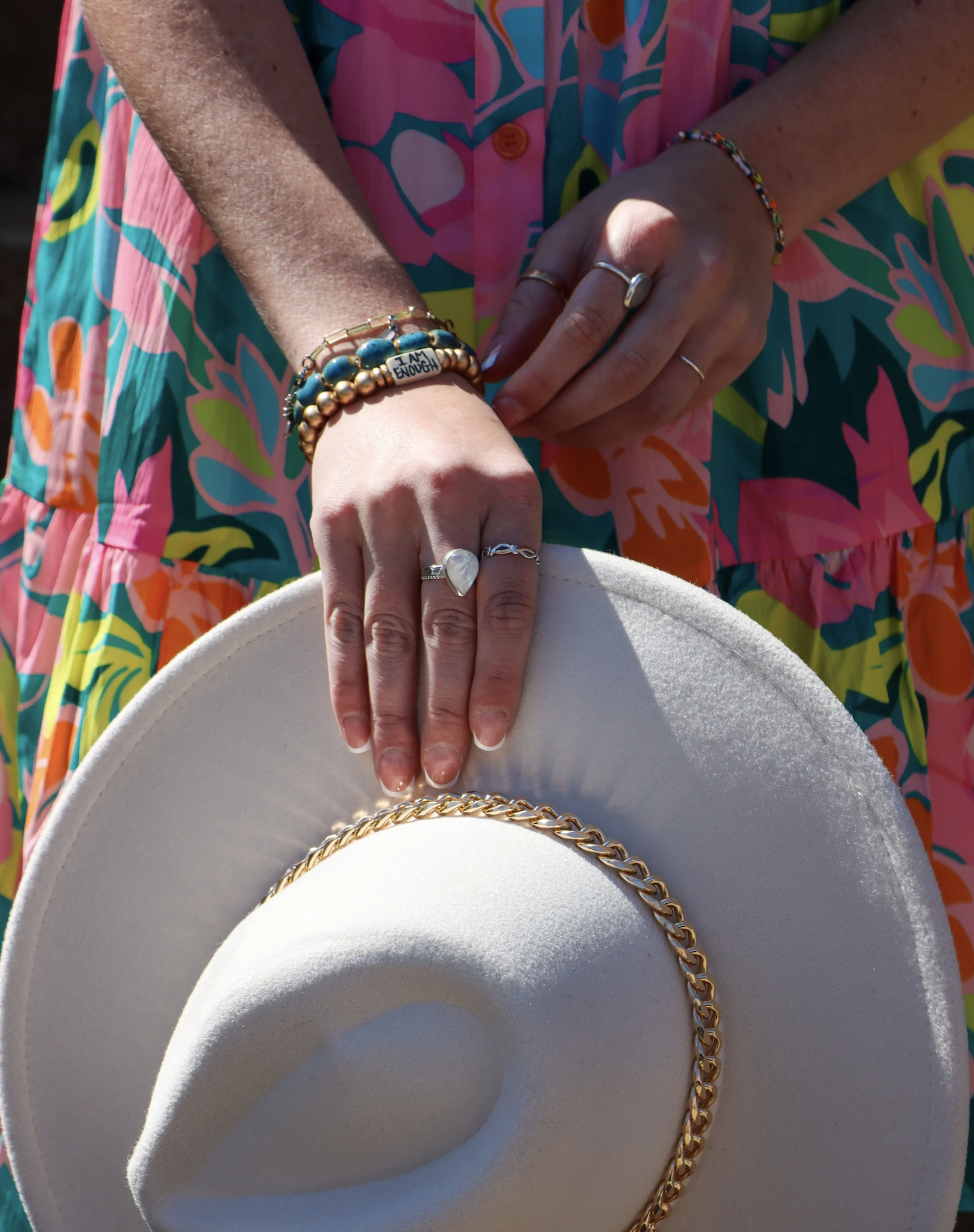 Close-up of a person's hand resting on a white hat with a gold chain, showing multiple jewelry pieces including rings and bracelets, against a colorful floral dress background.