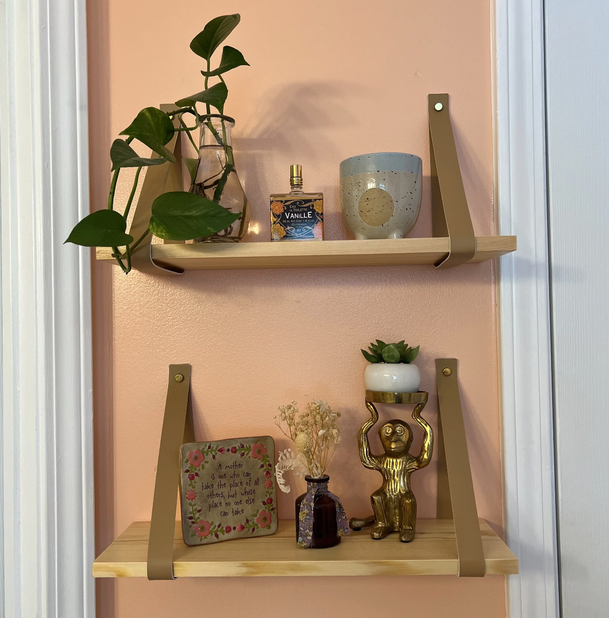 Decorative wall shelves with plants, a perfume bottle, a cup, a framed quote, dried flowers, and a small sculpture against a pink wall.