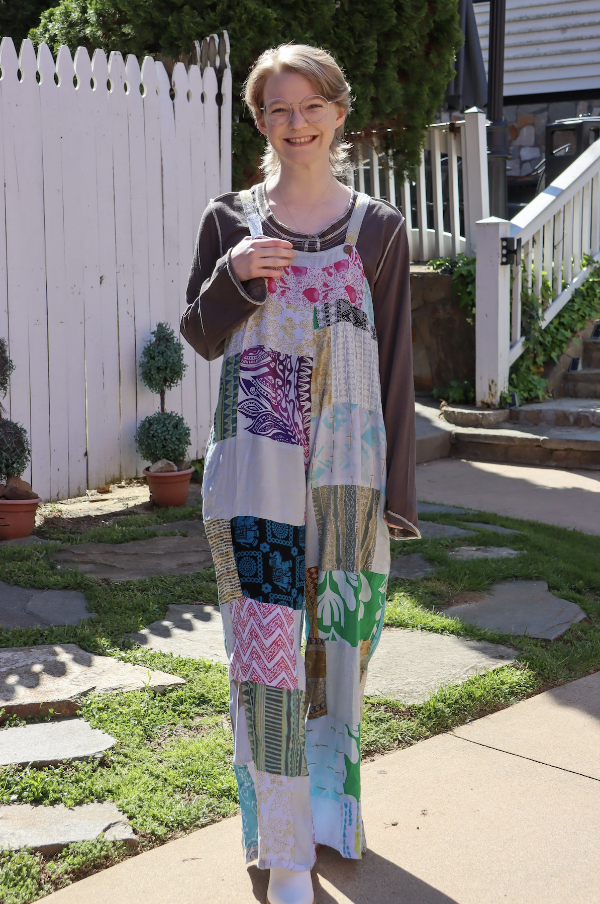 A young woman with glasses and short blonde hair smiling outside, wearing a long, colorful patchwork dress over a black long-sleeve shirt.