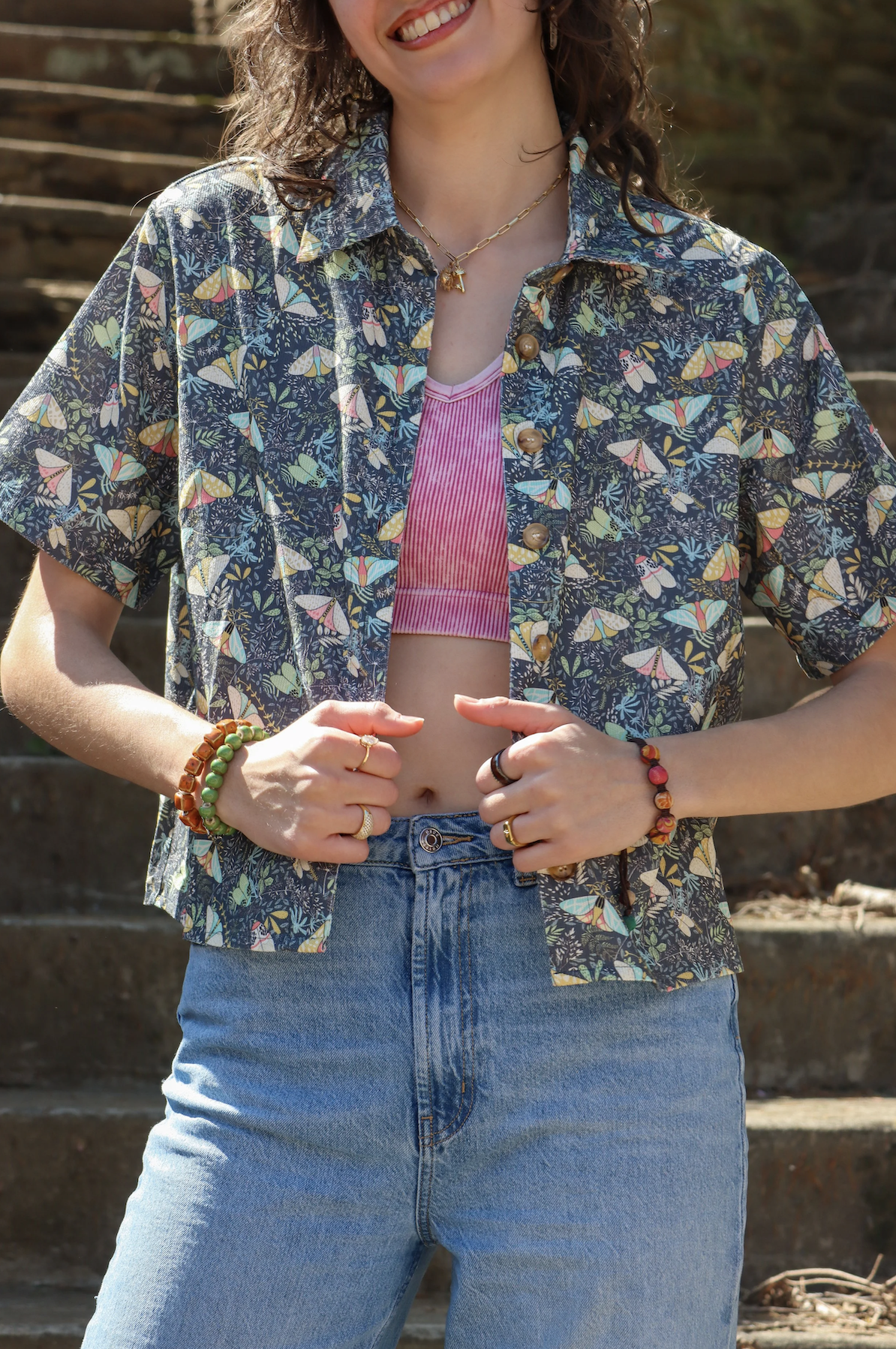 A smiling woman with curly hair wearing a patterned short-sleeve shirt over a pink striped crop top, jeans, and colorful jewelry, standing outdoors on stairs.