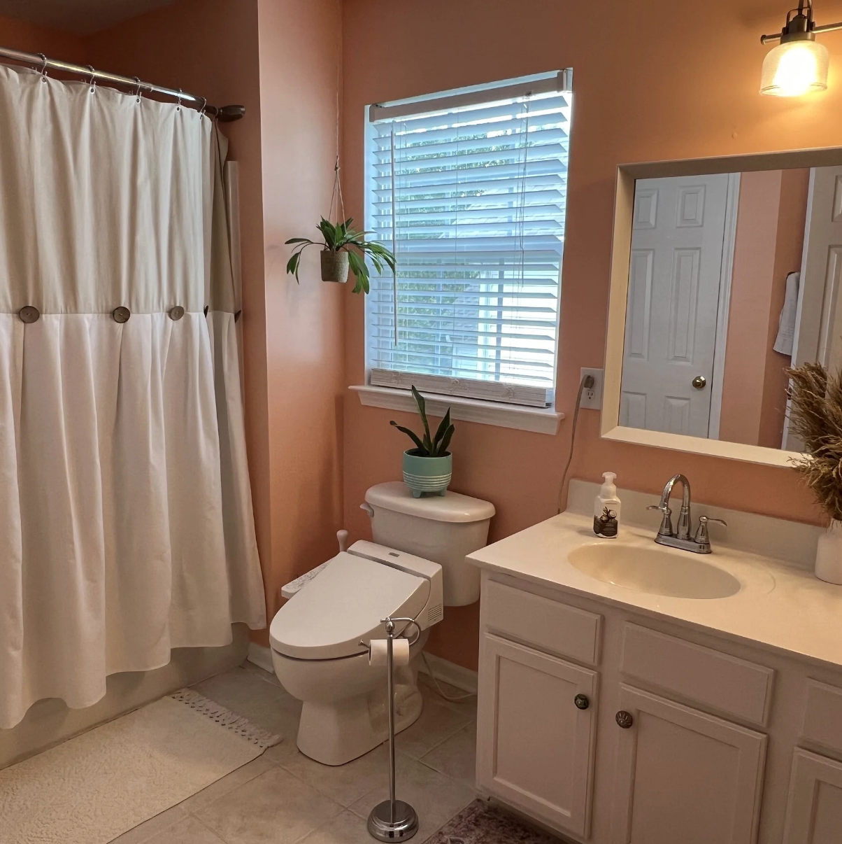 Bathroom with peach walls, white shower curtain, toilet, white vanity with sink, mirror, window with blinds, potted plants, and a light fixture.