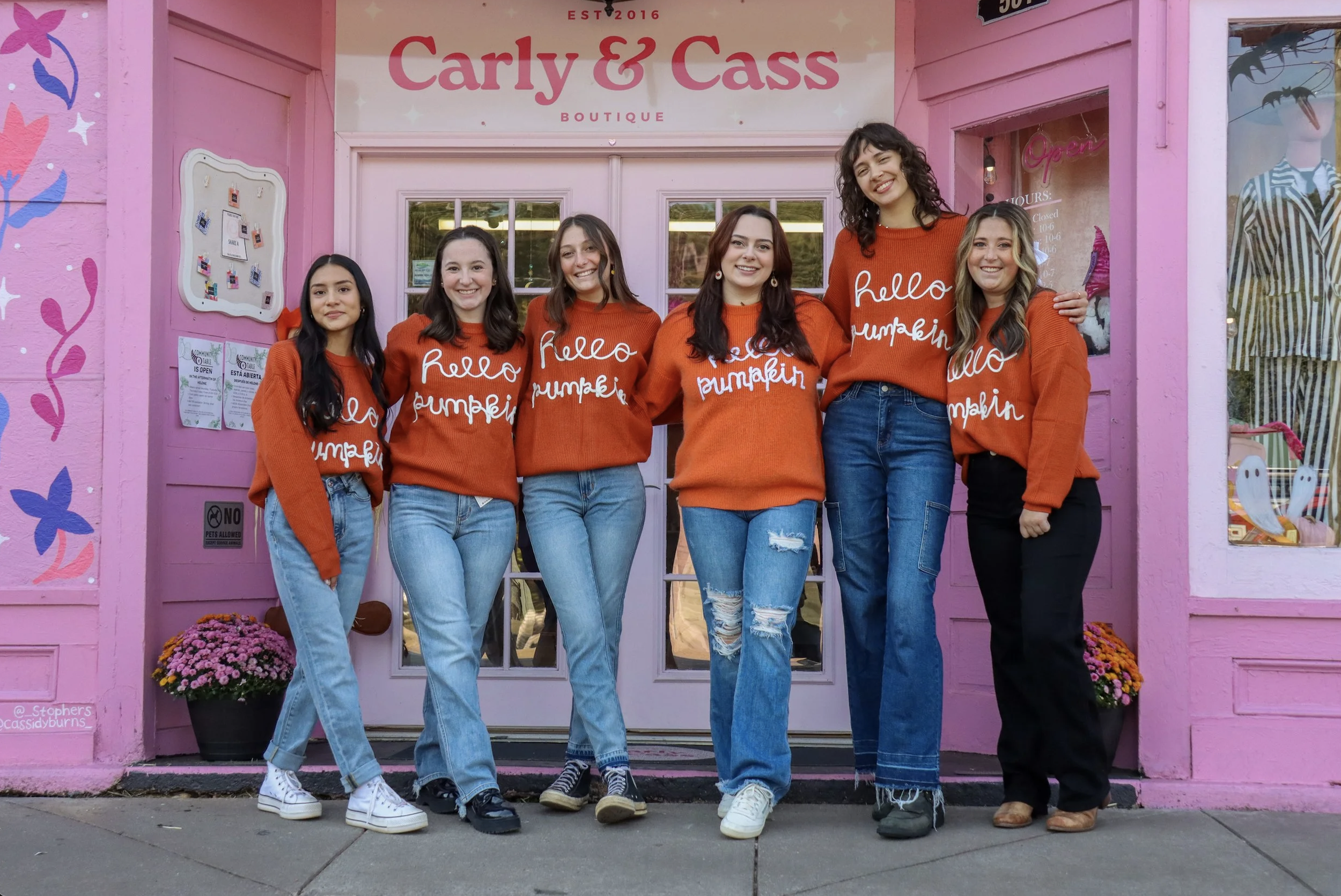 Group of six young women standing in front of a pink boutique store called Carly & Cass, all wearing matching orange sweaters with white text, and jeans, smiling.