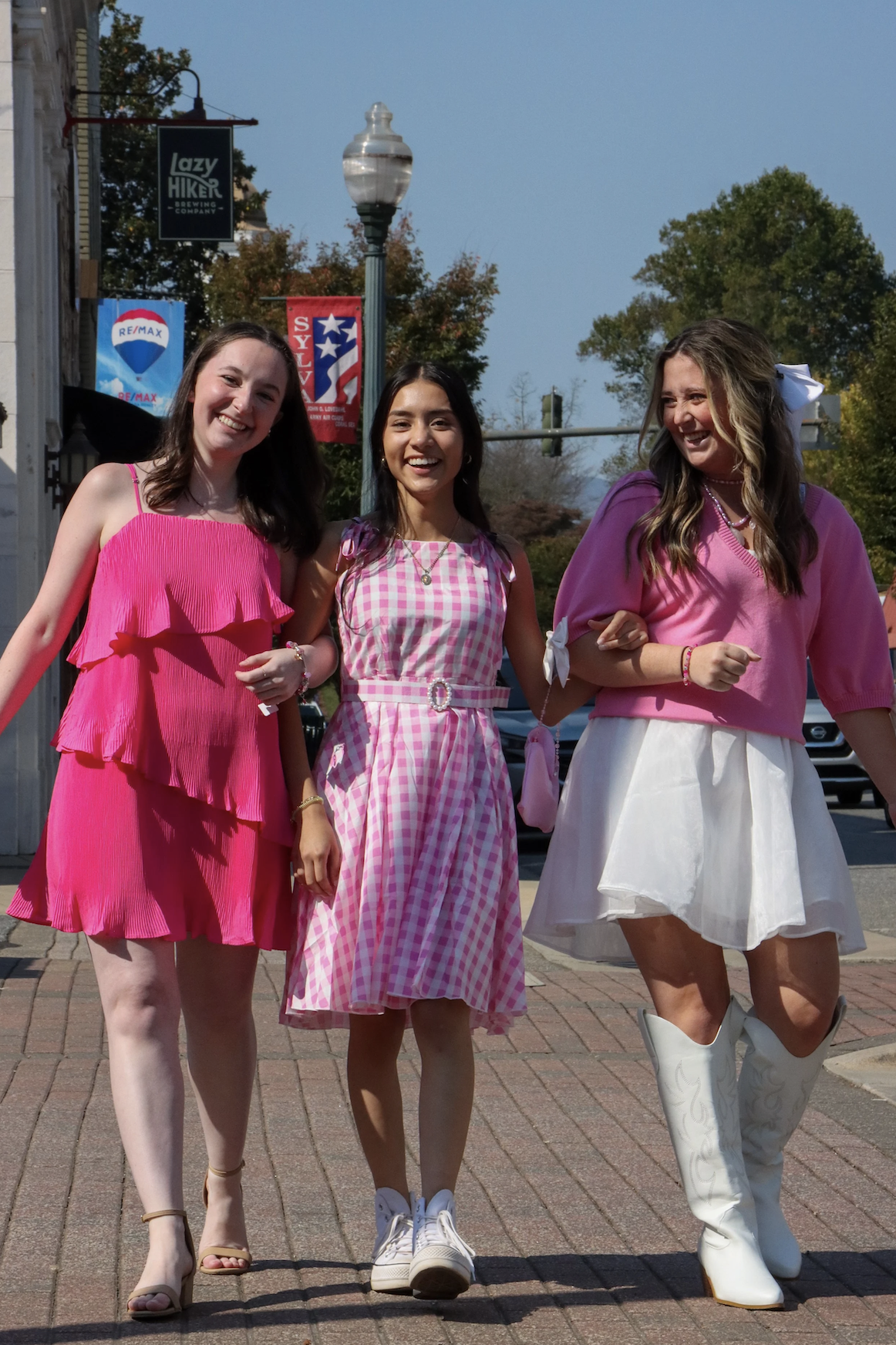 Three young women walking together outdoors, smiling, dressed in pink and white outfits, on a city street with flags, trees, and a lamp post in the background.