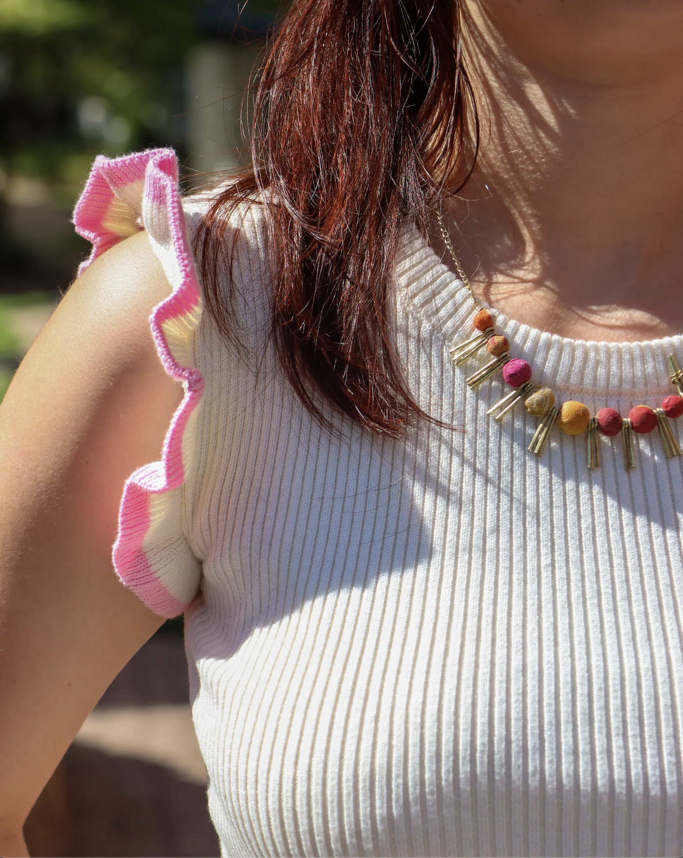 Close-up of a woman wearing a white ribbed sleeveless top with ruffled pink and white trim on the shoulder, and a colorful beaded necklace, outdoors with sunlight and greenery.