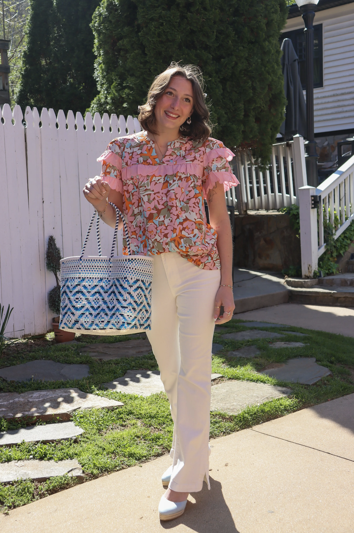 A woman with shoulder-length brown hair standing outside on a sunny day, wearing a floral blouse with pink ruffles, cream-colored pants, and white shoes, holding a woven tote bag with a blue and white pattern, in front of a white fence, trees, and a 