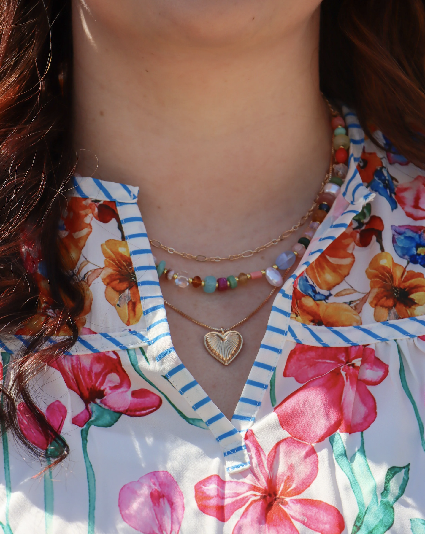 Close-up image of a woman wearing layered necklaces, including a gold chain with a heart-shaped pendant, and a colorful floral shirt with a collar, open at the top.