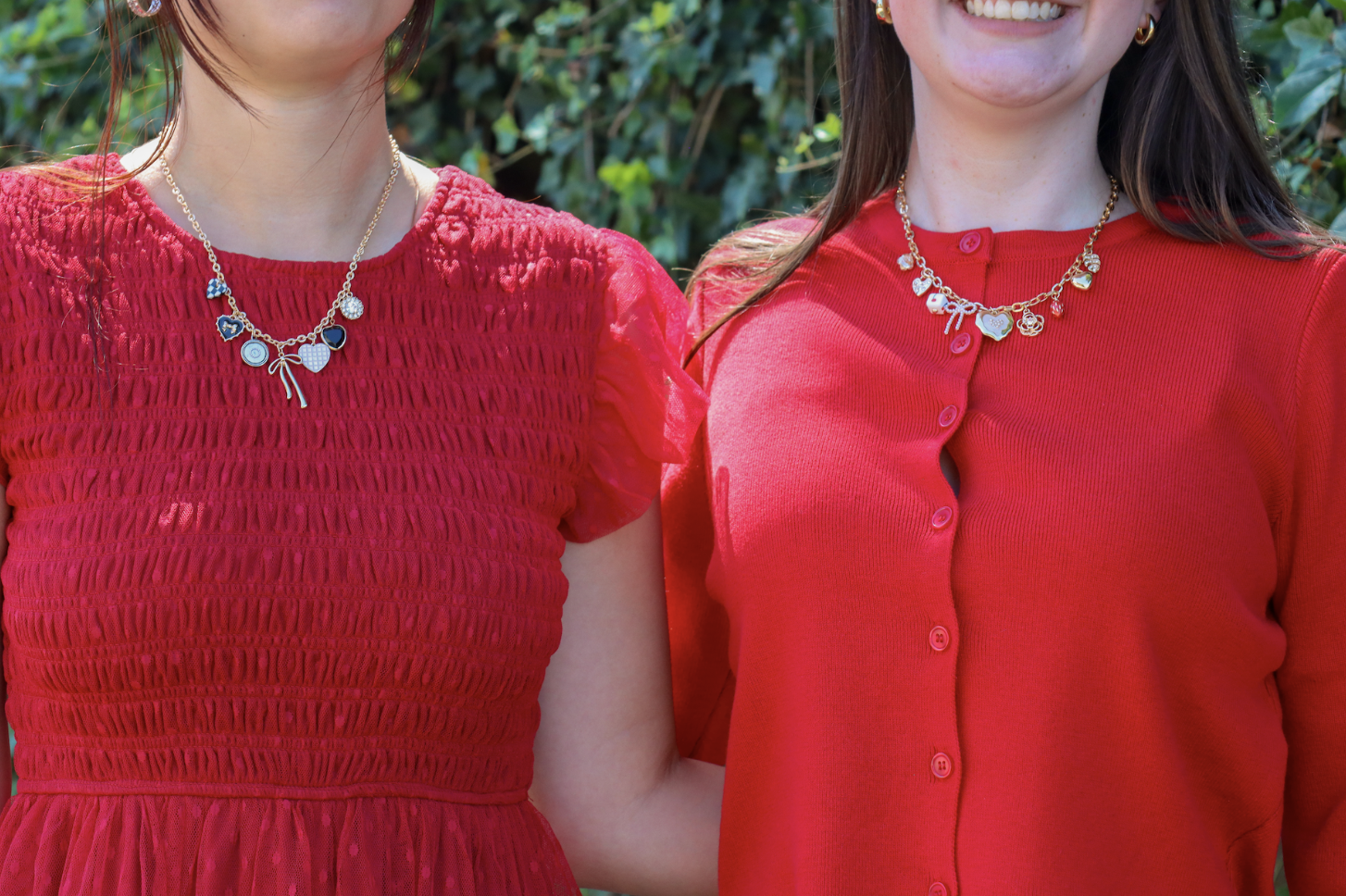 Two women wearing red dresses and necklaces, standing outdoors with greenery.