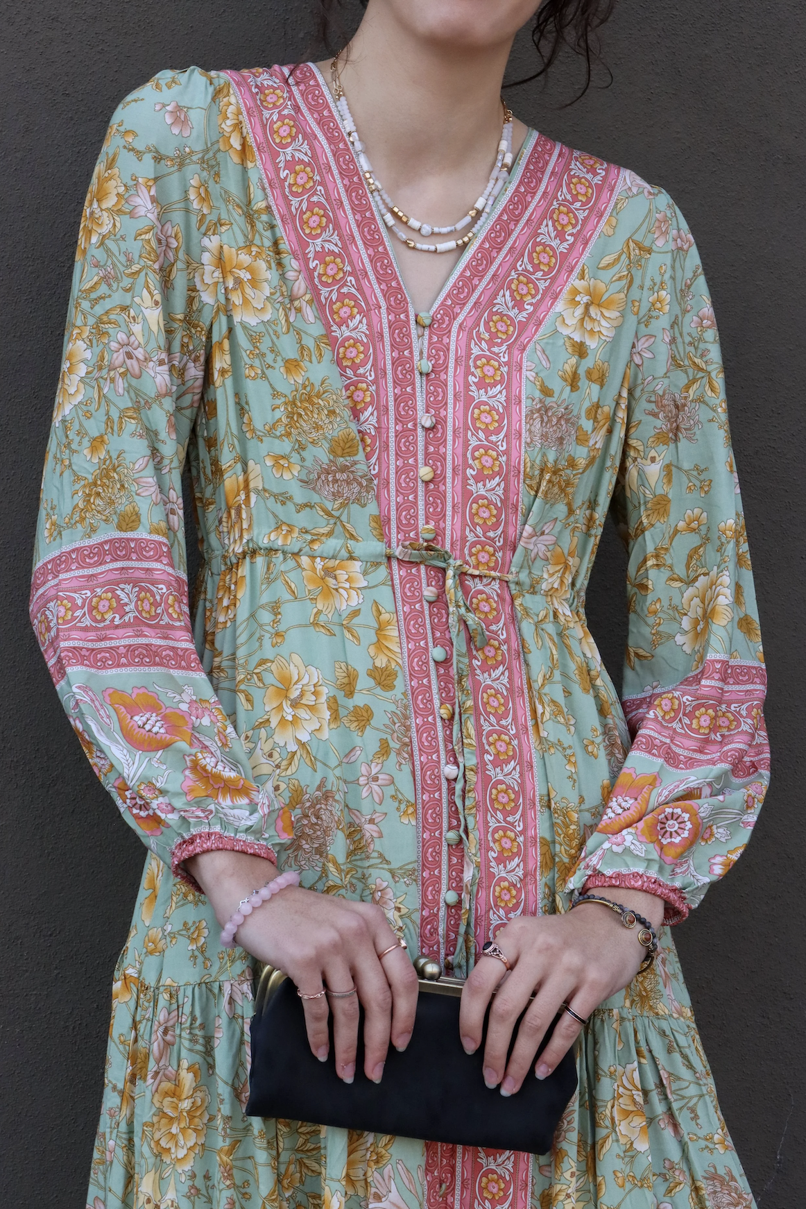 A woman wearing a long, floral dress with pink and yellow accents, holding a small black clutch, and accessorized with necklaces, bracelets, and rings, standing against a dark gray wall.