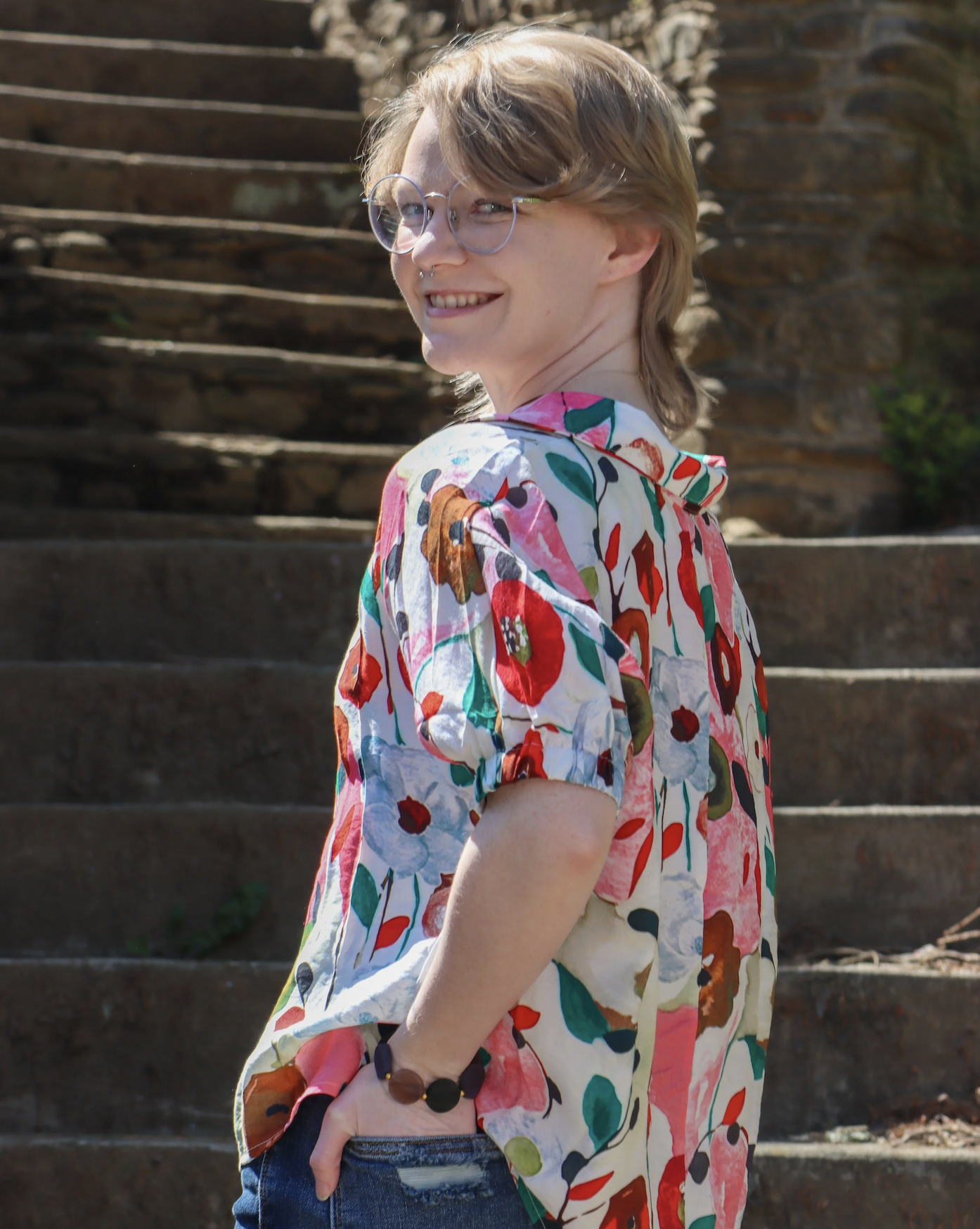 A young woman with short blond hair, glasses, and a septum piercing, smiling and looking back over her shoulder, wearing a colorful floral shirt and ripped jeans, standing outdoors near stone stairs.