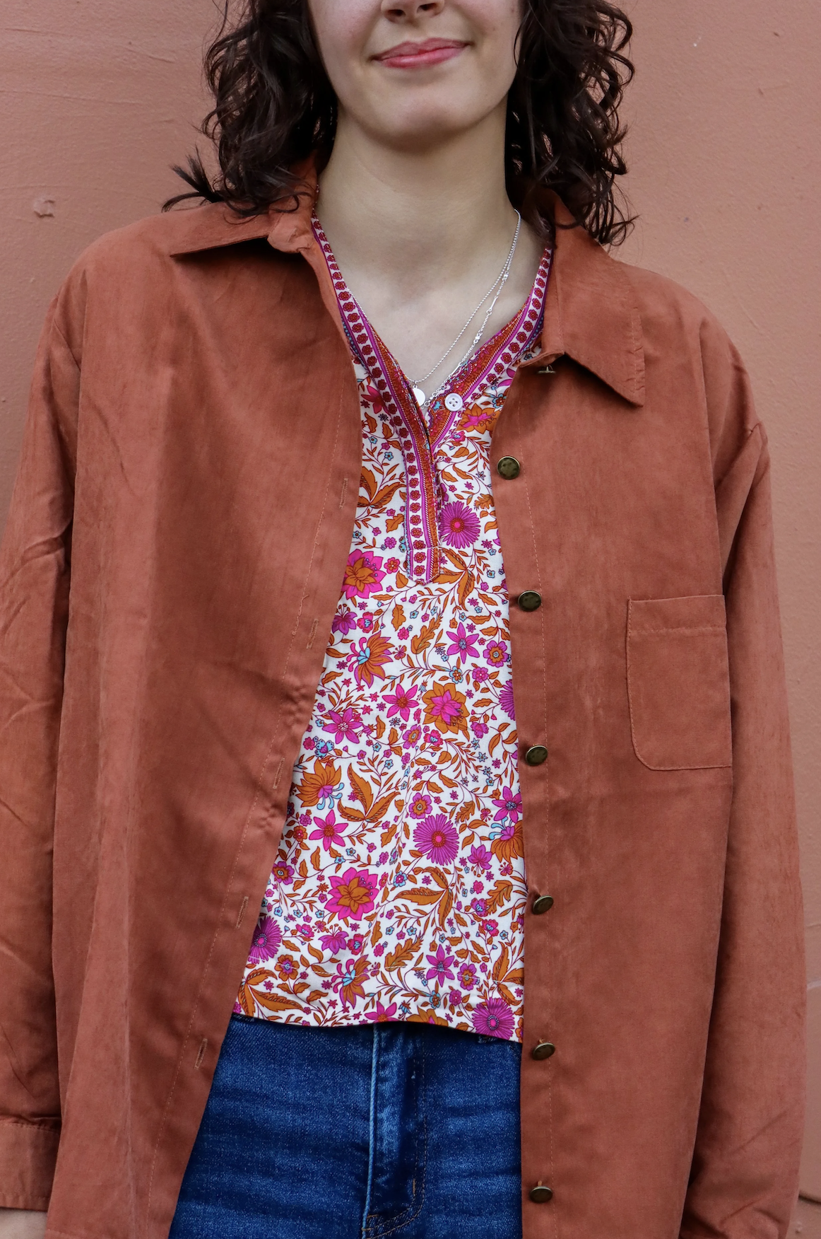 A woman wearing an orange-brown jacket over a colorful floral blouse, standing against a pink wall.
