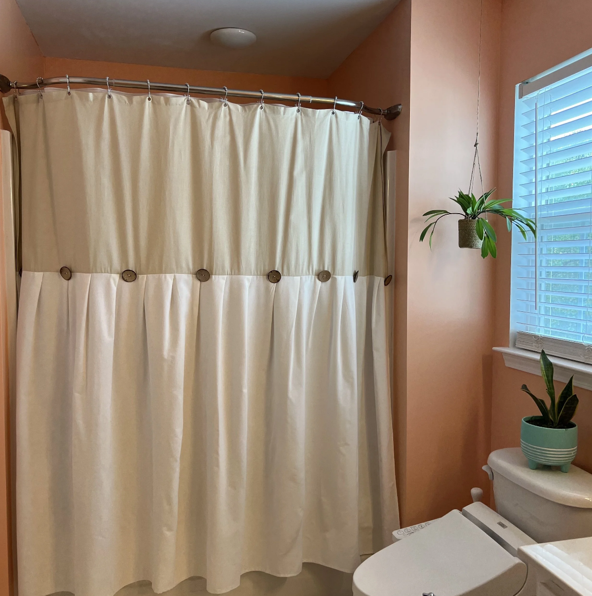 Bathroom with peach-colored walls, a window with blinds, a white shower curtain, and plants on the window sill and toilet tank.