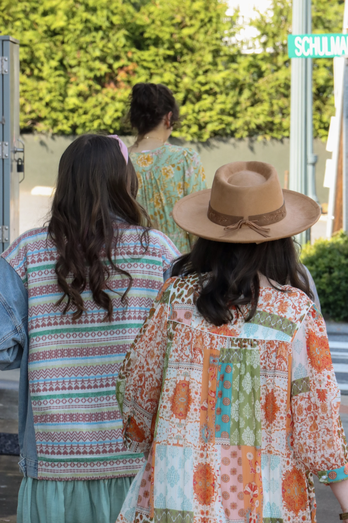 Three women standing outdoors near a street sign that reads 'Schulman.' They are dressed in colorful, patterned clothing and are viewed from behind. The woman in the foreground is wearing a hat and a patchwork-patterned dress, and the woman to her le