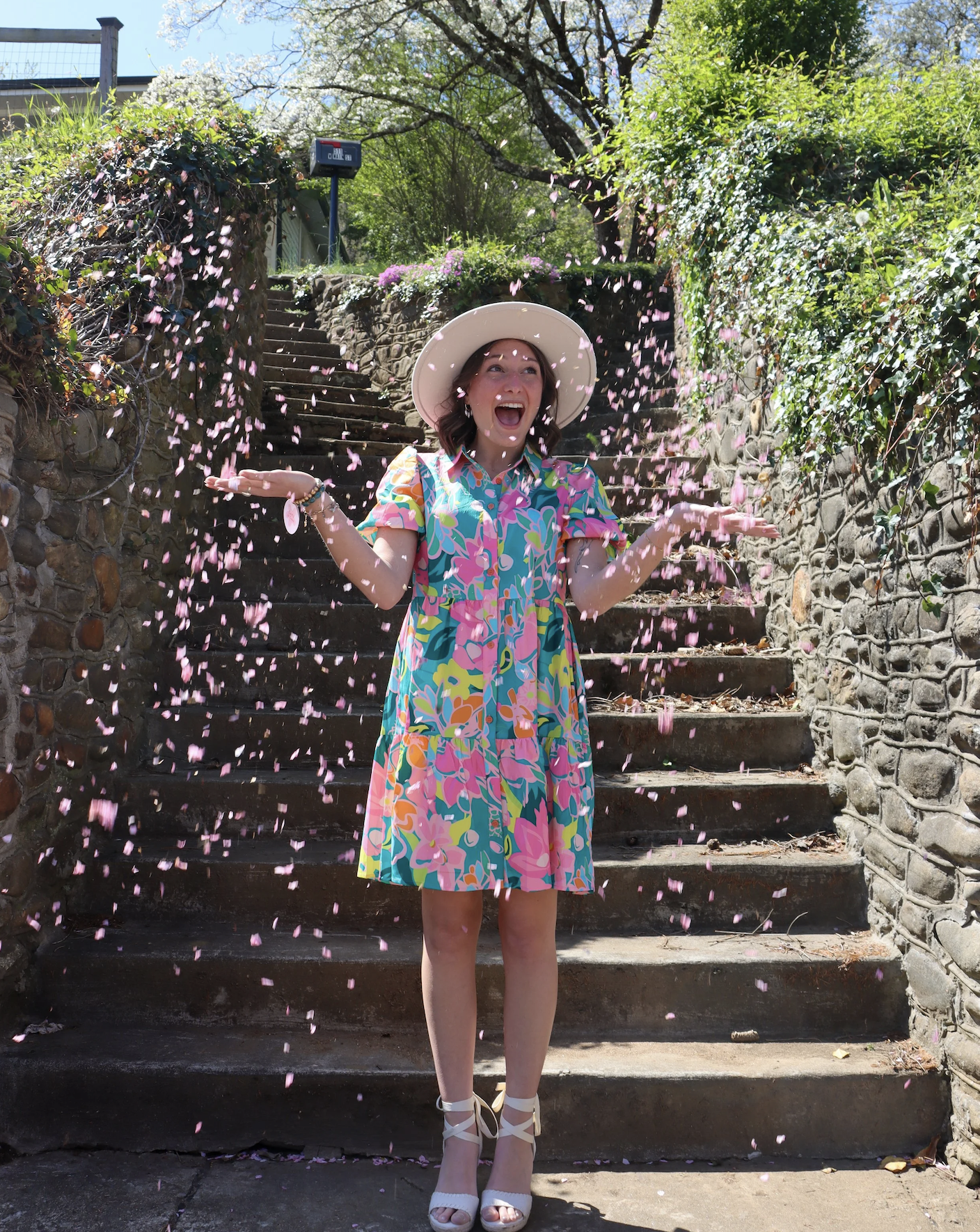 A woman in a colorful floral dress and white hat celebrating on stone steps with falling pink flower petals.