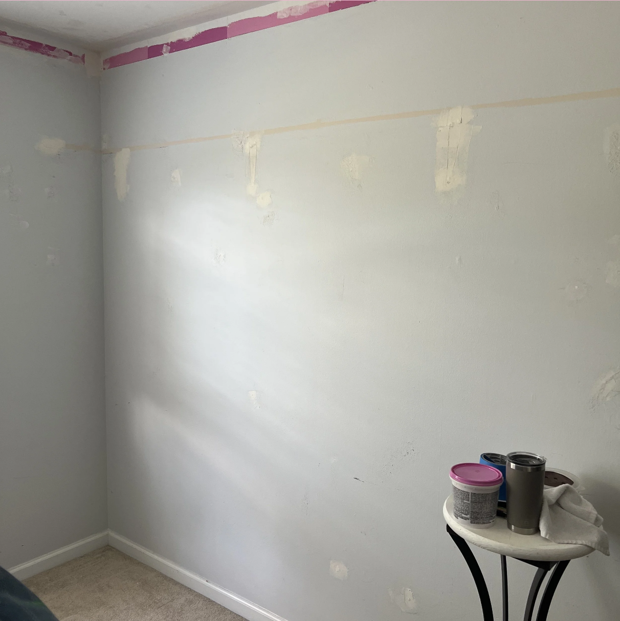 Interior wall undergoing painting or renovation with patches and masking tape near the ceiling, a small table with paint cans, a tumbler, and a cloth in the corner.