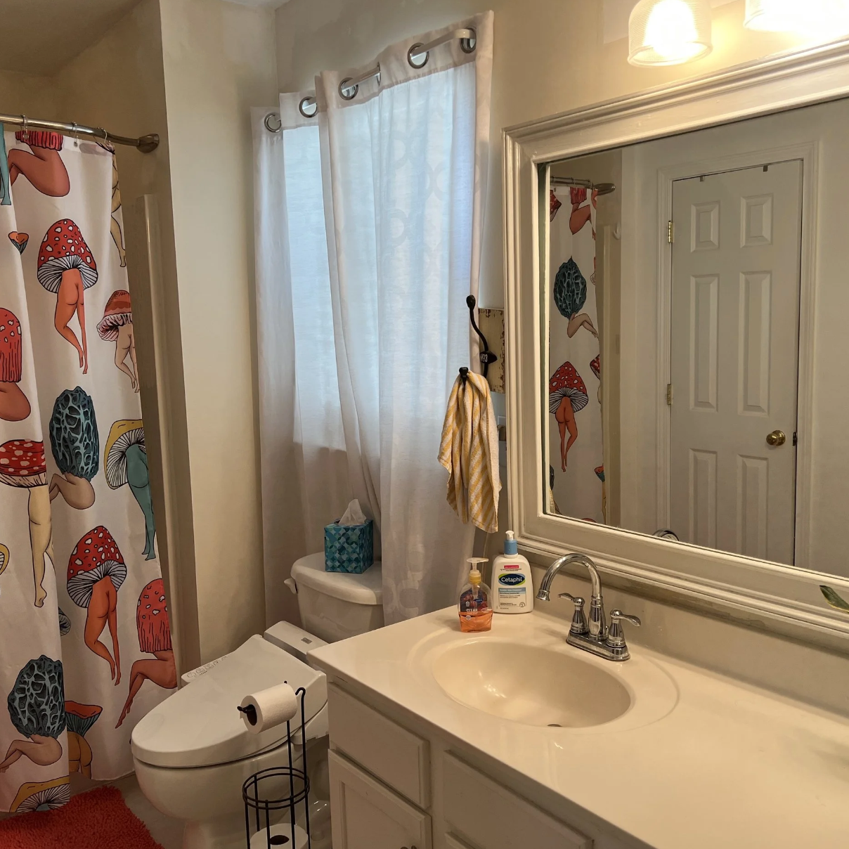 A bathroom with a shower curtain featuring colorful mushroom and fairy illustrations. There is a white toilet with a box of tissues and a yellow striped towel hanging on a hook. The sink area has hand soap, sanitizer, and a mirror above it. Light ent