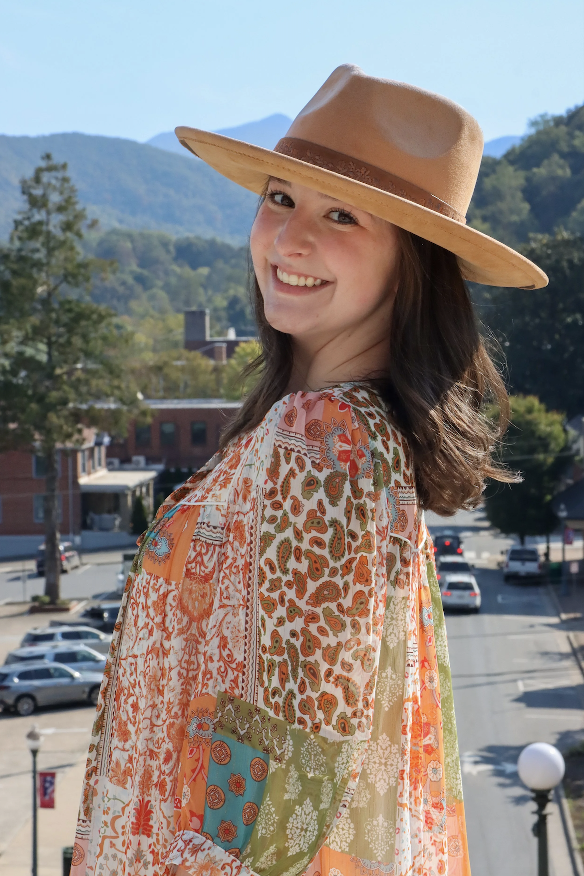 A young woman with long brown hair smiling, wearing a wide-brimmed tan hat and a colorful bohemian-style dress, standing outdoors with a mountain range and cityscape in the background.