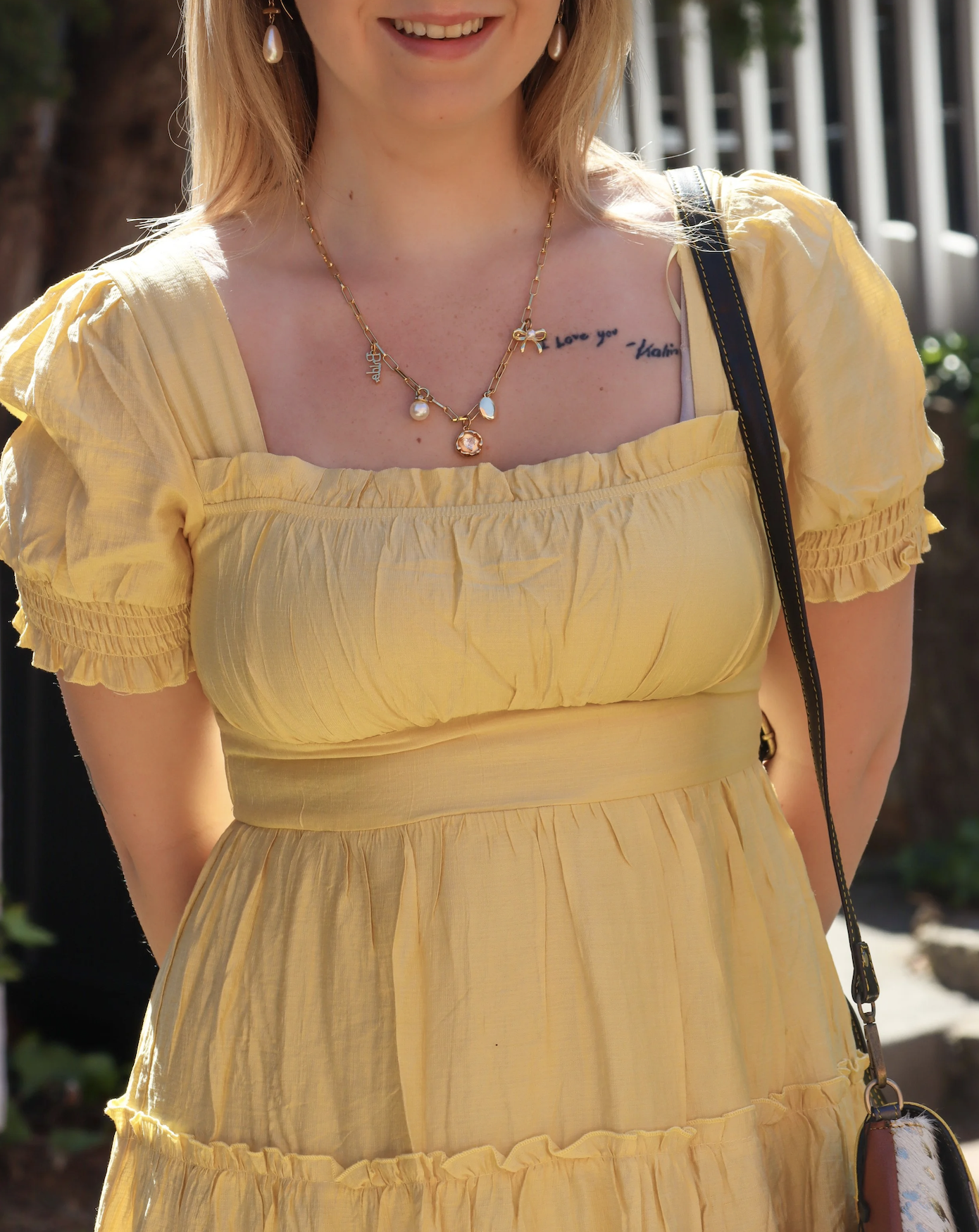A woman wearing a yellow dress with puff sleeves and a gathered waist, smiling outdoors. She has a necklace with various charms, pearl earrings, and a black strap shoulder bag.
