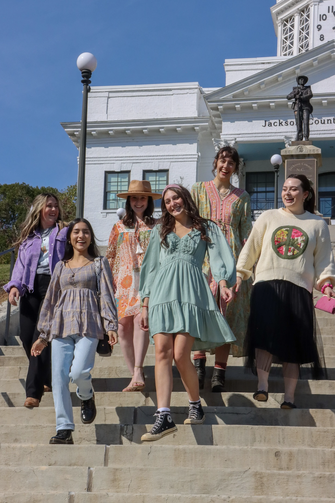 A group of seven women, smiling and walking down the steps of a white courthouse with the sign "Jackson County". They are dressed in colorful, casual clothing and the weather is sunny and clear.