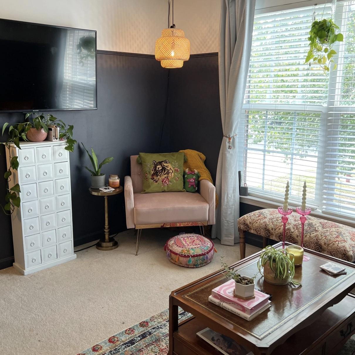 A cozy living room with a pink armchair decorated with a green panda pillow, a yellow blanket, and a tiger cushion. There is a small round side table with candles and a potted plant next to the armchair. Large windows with white blinds and curtains l