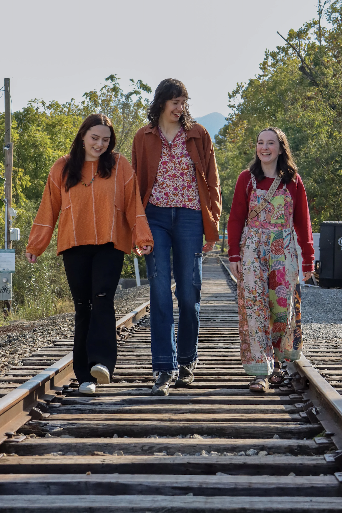Three young women walking on a railroad track, smiling and enjoying each other's company, with trees and a mountain in the background.