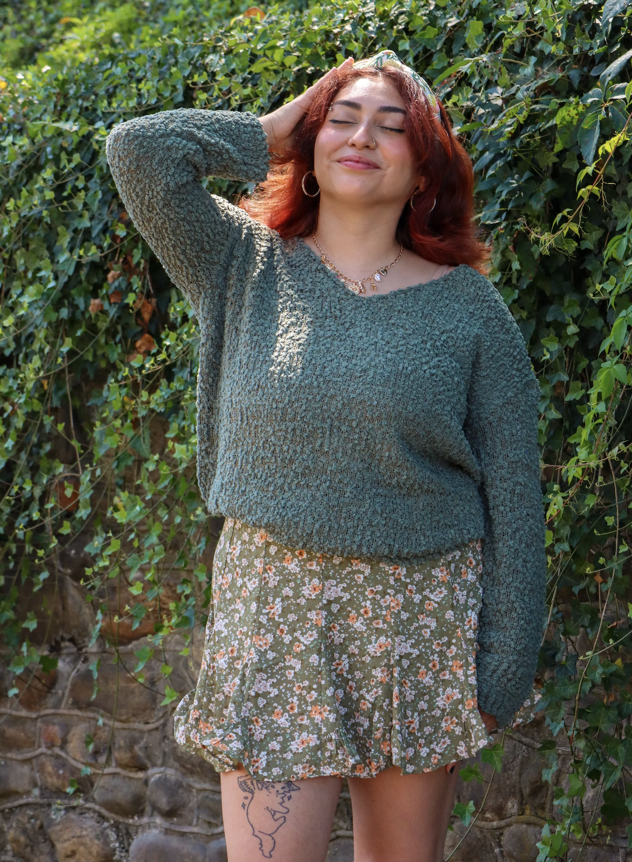A young woman with red hair, closed eyes, and a gentle smile stands outdoors against green leafy bushes. She is wearing a textured green sweater, floral patterned shorts, hoop earrings, a necklace, and a nose ring, with a visible tattoo of a face on 