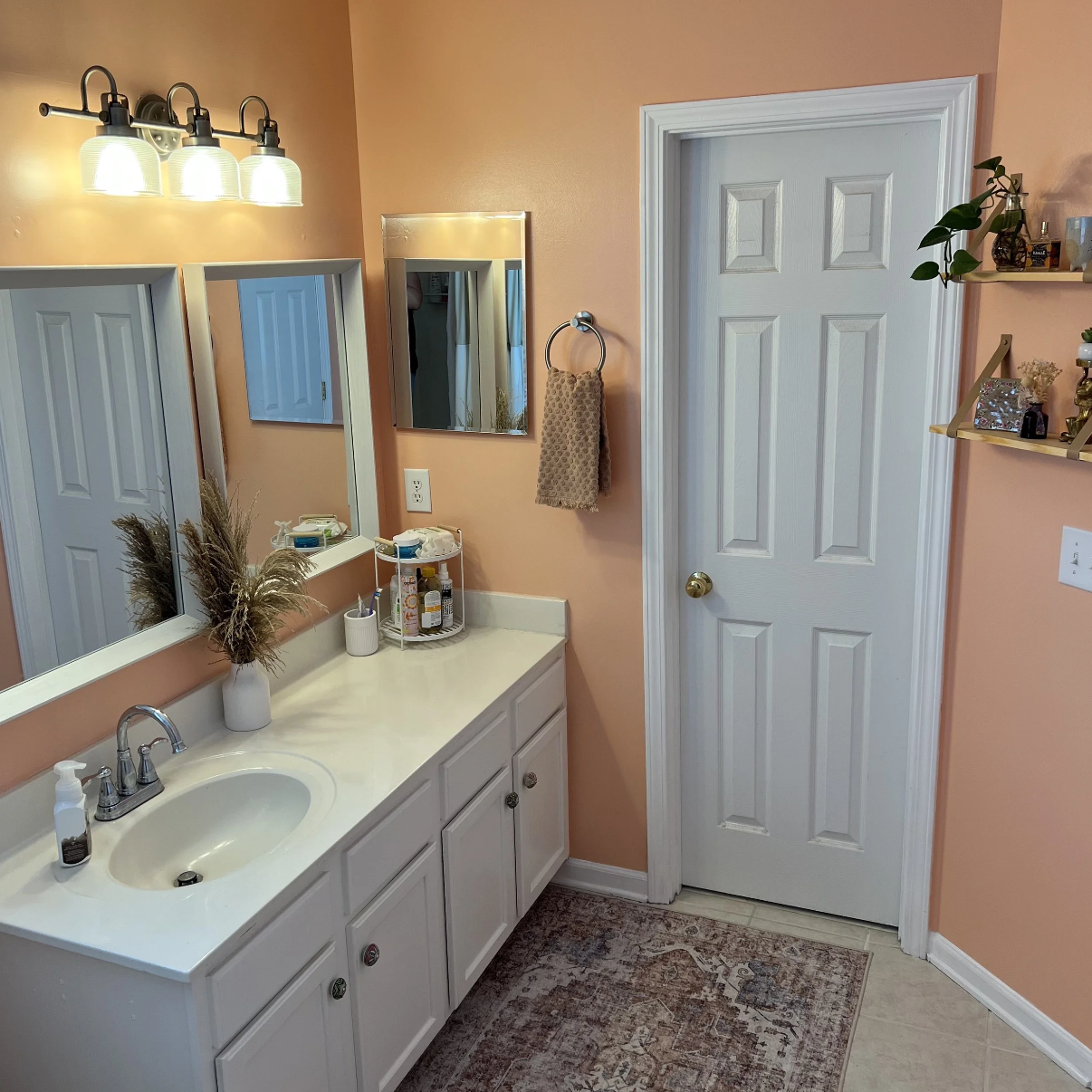 Bathroom with peach walls, white vanity with a sink, mirror, and shelves, decorated with dried plants, and a door on the right, with a small rug on the floor.