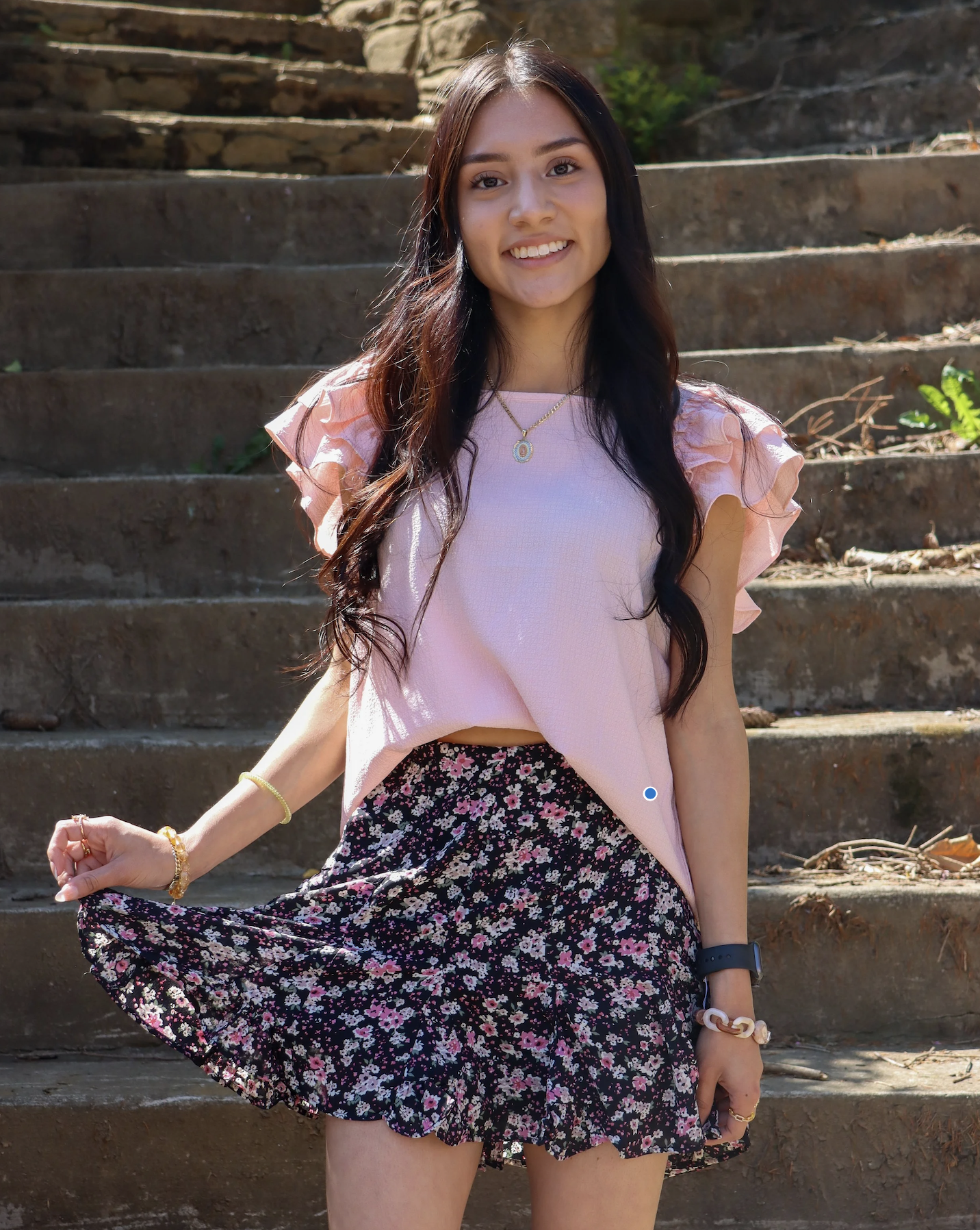 A young woman with long dark hair, smiling, standing outdoors on stone steps, wearing a pink top, floral skirt, and jewelry.