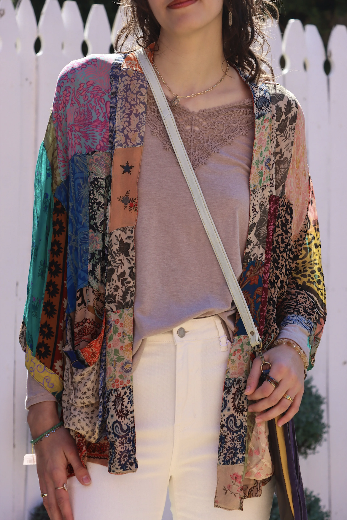 A woman is wearing a multicolored patchwork kimono over a light pink lace-trimmed top, paired with white pants. She has jewelry including rings, a bracelet, and a gold chain necklace. She is standing outdoors with a white picket fence in the backgrou