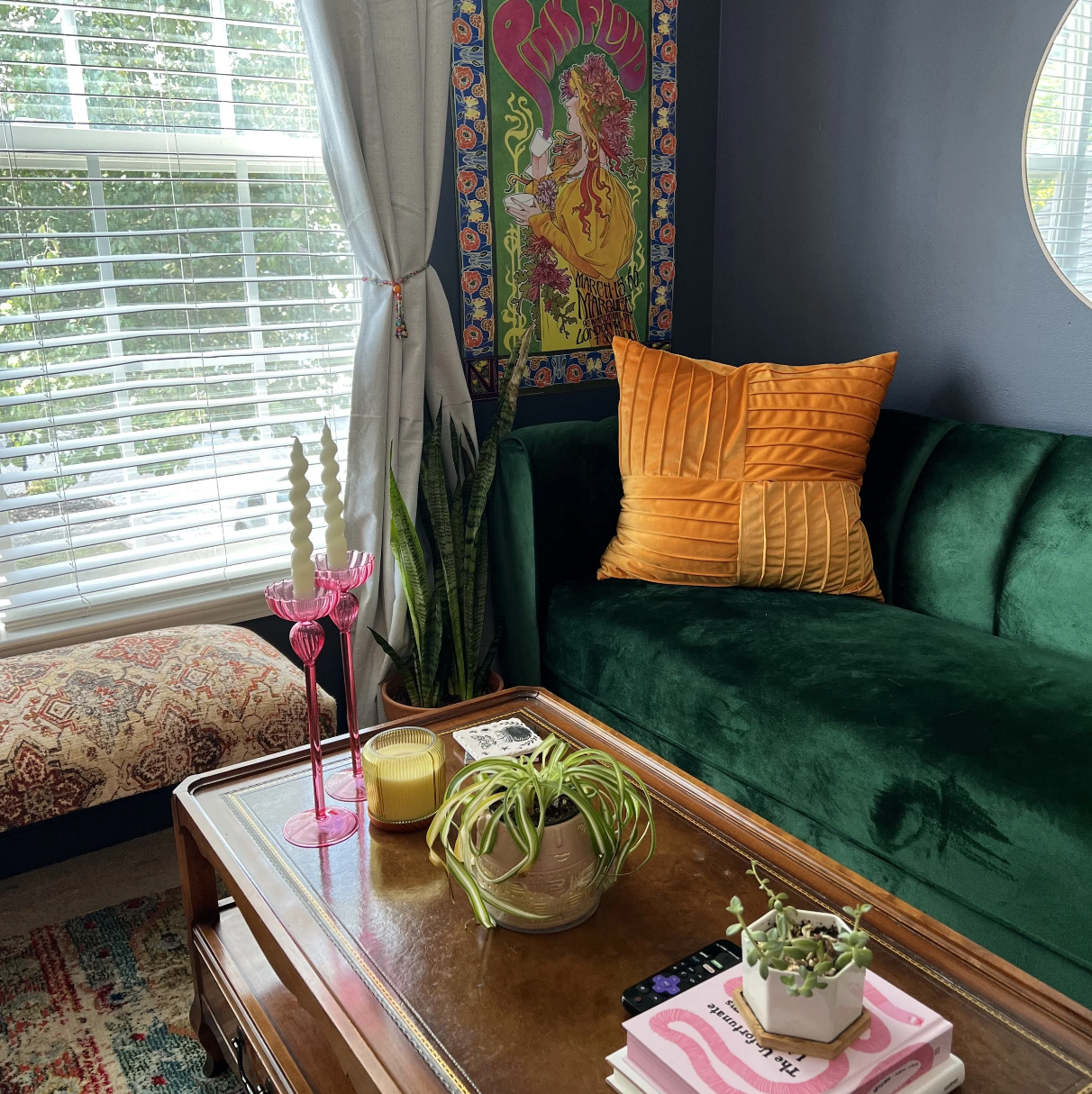 A cozy living room corner with a green velvet sofa, decorated with large orange pillows. A wooden coffee table holds potted plants, candles, and a remote control. Behind the sofa is a colorful tapestry and a window with white blinds and drapes. The w
