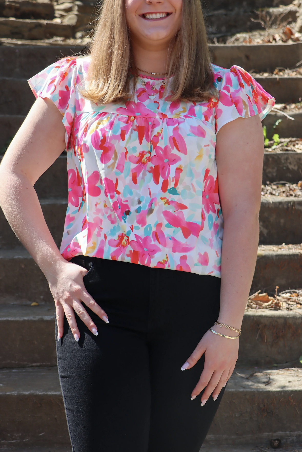 A woman is standing outdoors on a set of stone steps, wearing a colorful floral top and black pants, with her left hand on her hip, smiling.