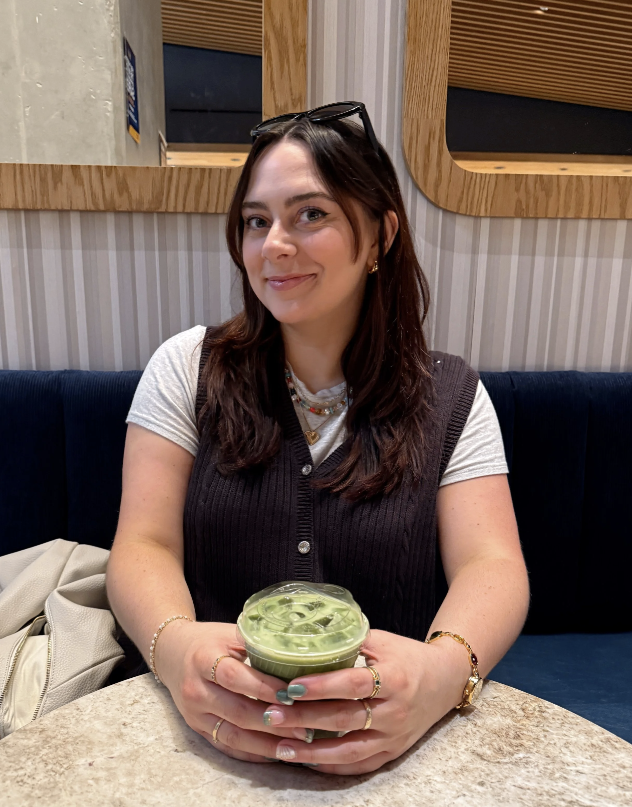 A young woman with dark brown hair and glasses on her head, smiling and holding a cup of matcha latte with ice, sitting at a table in a cafe.