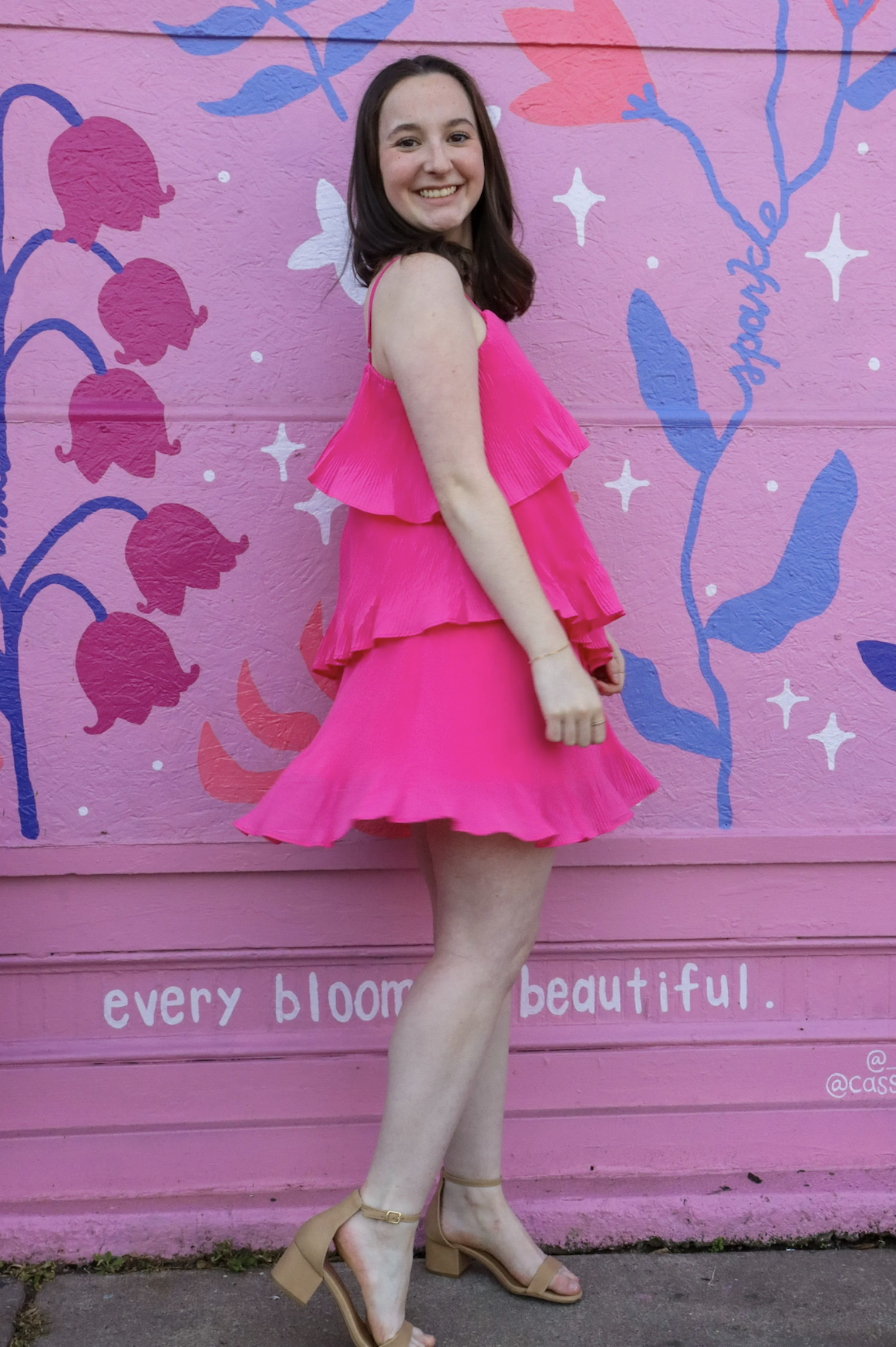 A woman with shoulder-length brown hair, smiling, wearing a bright pink ruffled dress and beige high-heeled sandals, standing against a pink wall with purple and white floral mural and the words "every bloom is beautiful" written on it.