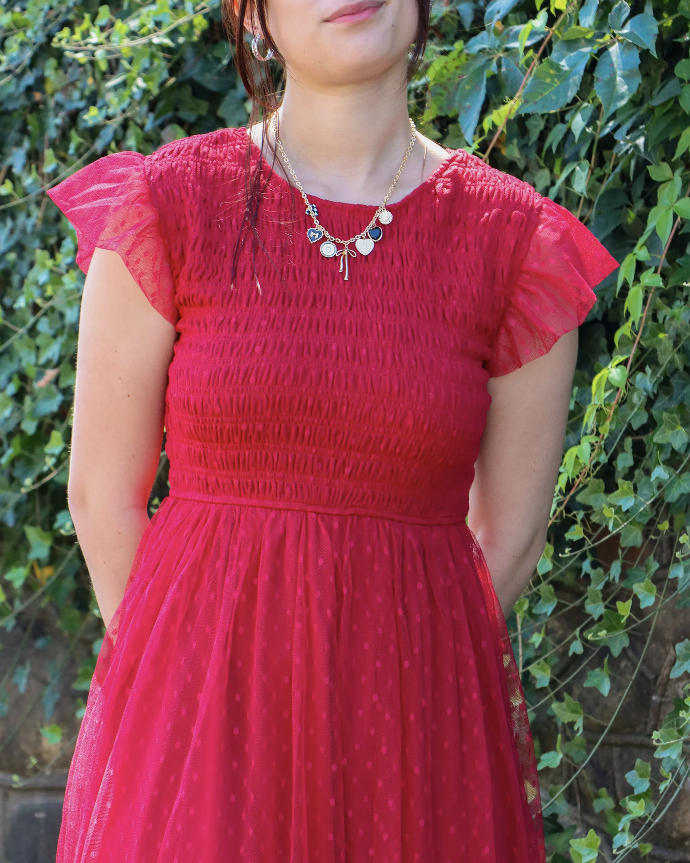 Close-up of a woman wearing a red dress with crinkled fabric and ruffled short sleeves, standing outdoors in front of green leafy plants, with a gold necklace featuring charms.