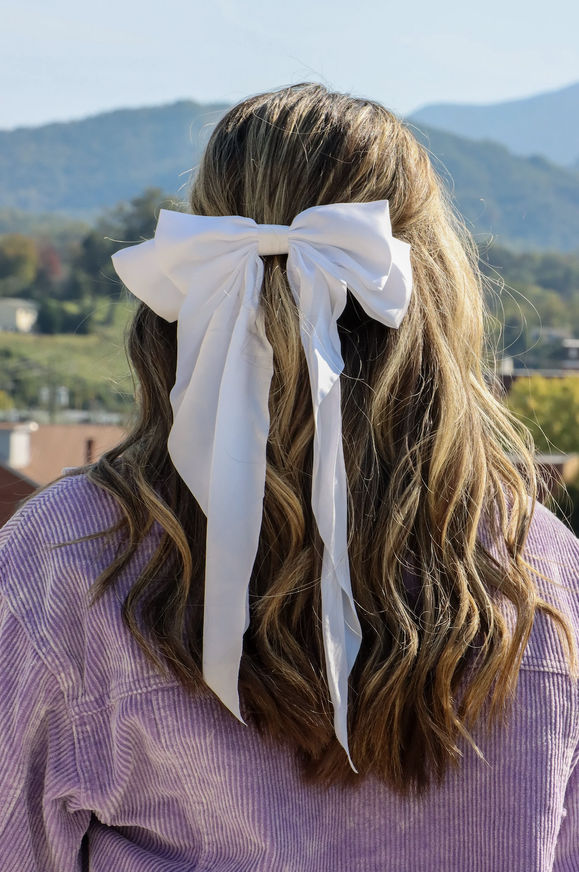 A person with long, wavy hair wearing a large white bow in their hair, standing outdoors with mountains in the background.