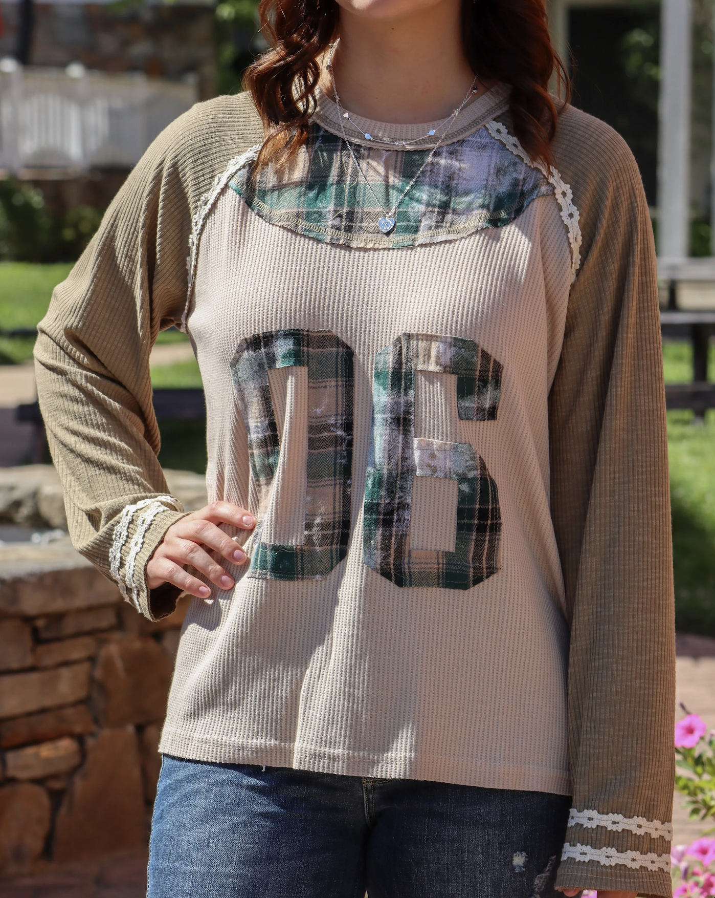 Person wearing a long-sleeve beige and plaid shirt with the number 03 on the front, standing outdoors with a brick wall and green foliage in the background.
