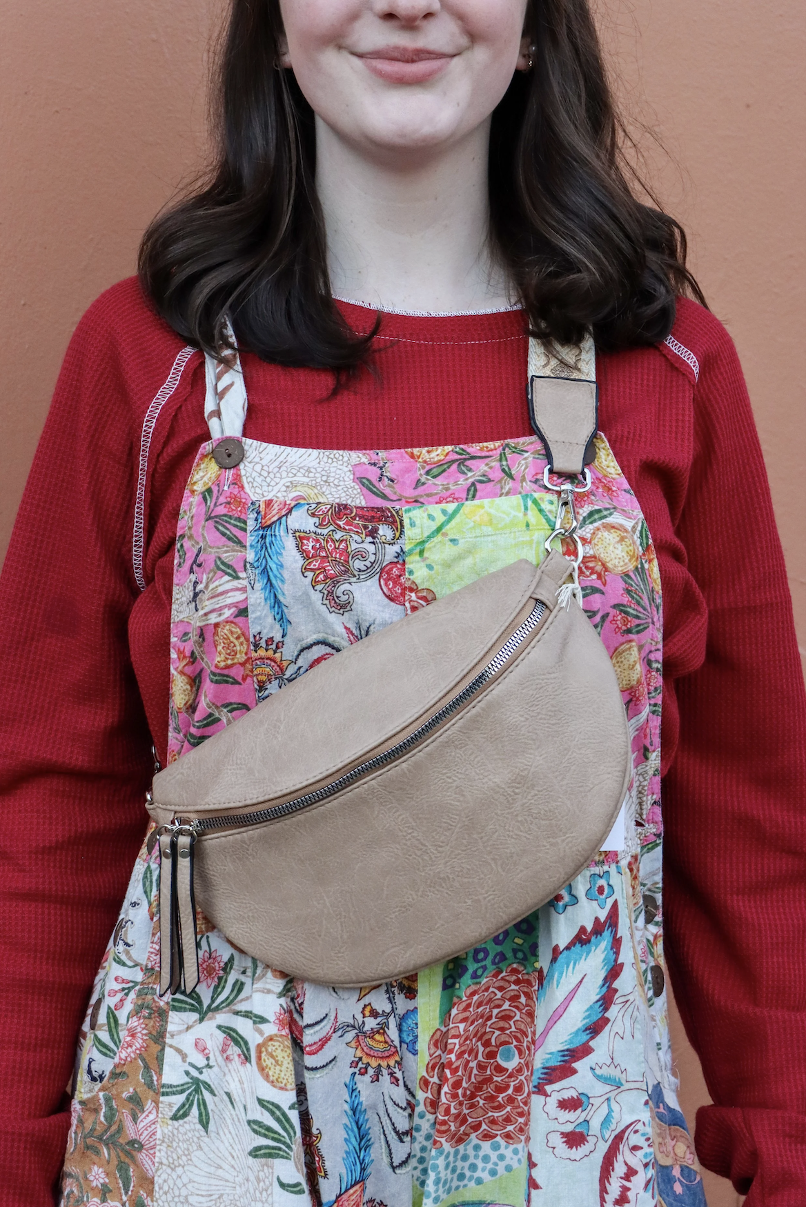 A person wearing a red long-sleeve shirt, a colorful patchwork apron, and a beige crossbody bag standing against a peach-colored wall.