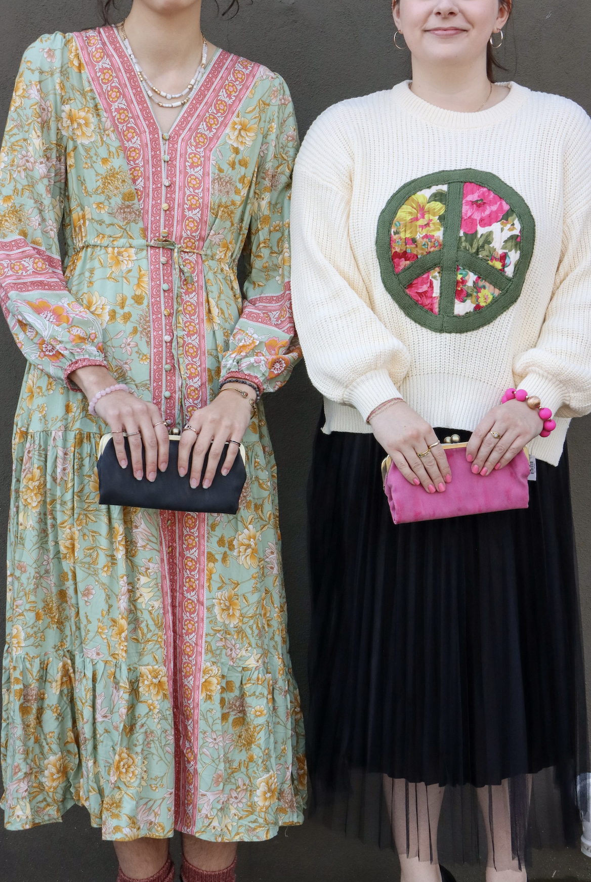Two women standing side by side, dressed in colorful outfits, holding small clutches, against a dark background.