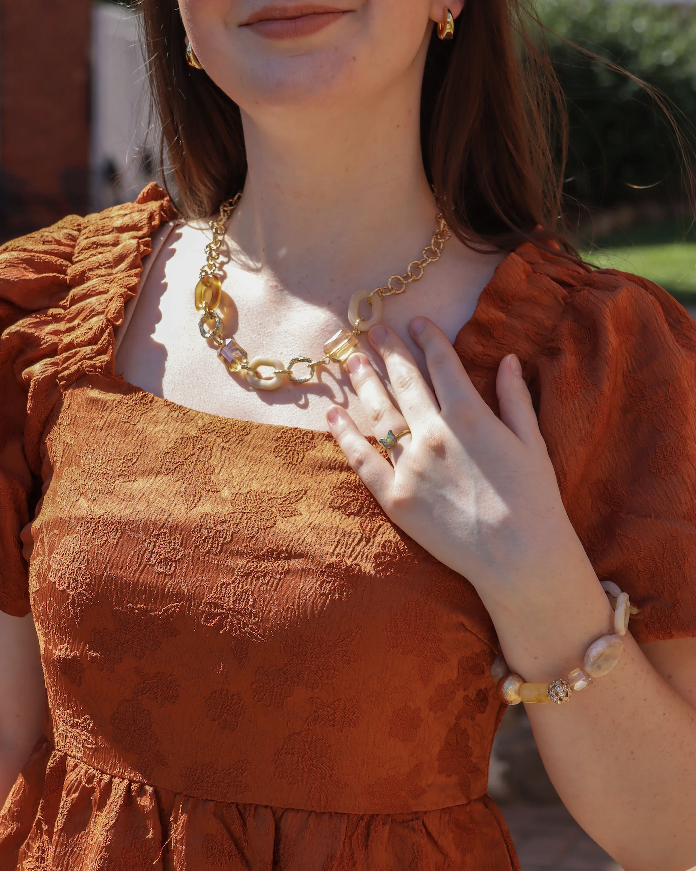 Close-up of a woman wearing a textured orange dress with jewelry, including a gold chain necklace, earrings, a ring, and a bracelet with large beads, outdoors