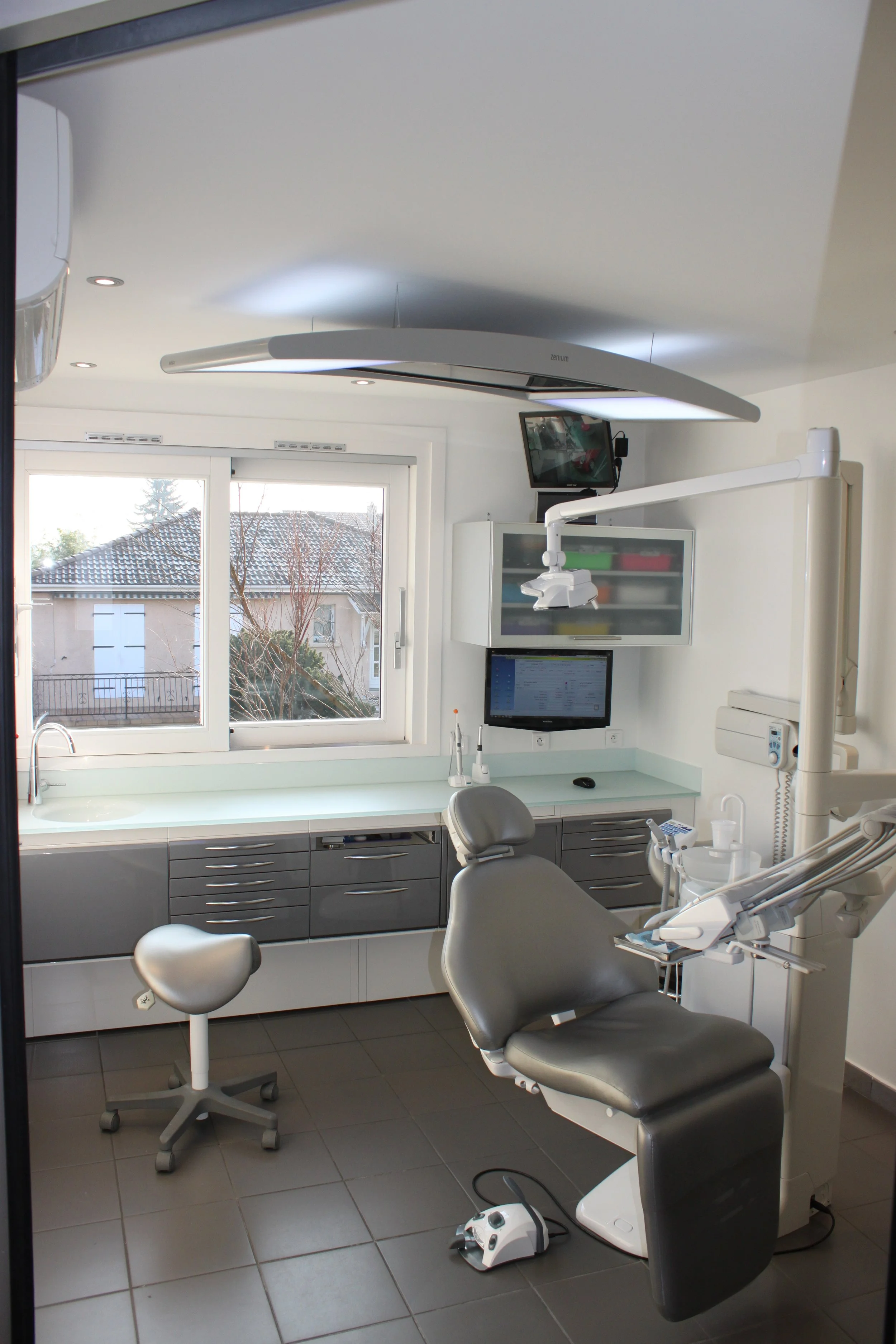 Salle de soins dentaires équipée d'une chaise de dentiste, d'un ordinateur, d'outils dentaires, d'un éclairage et d'une fenêtre donnant sur un toit et des arbres.
