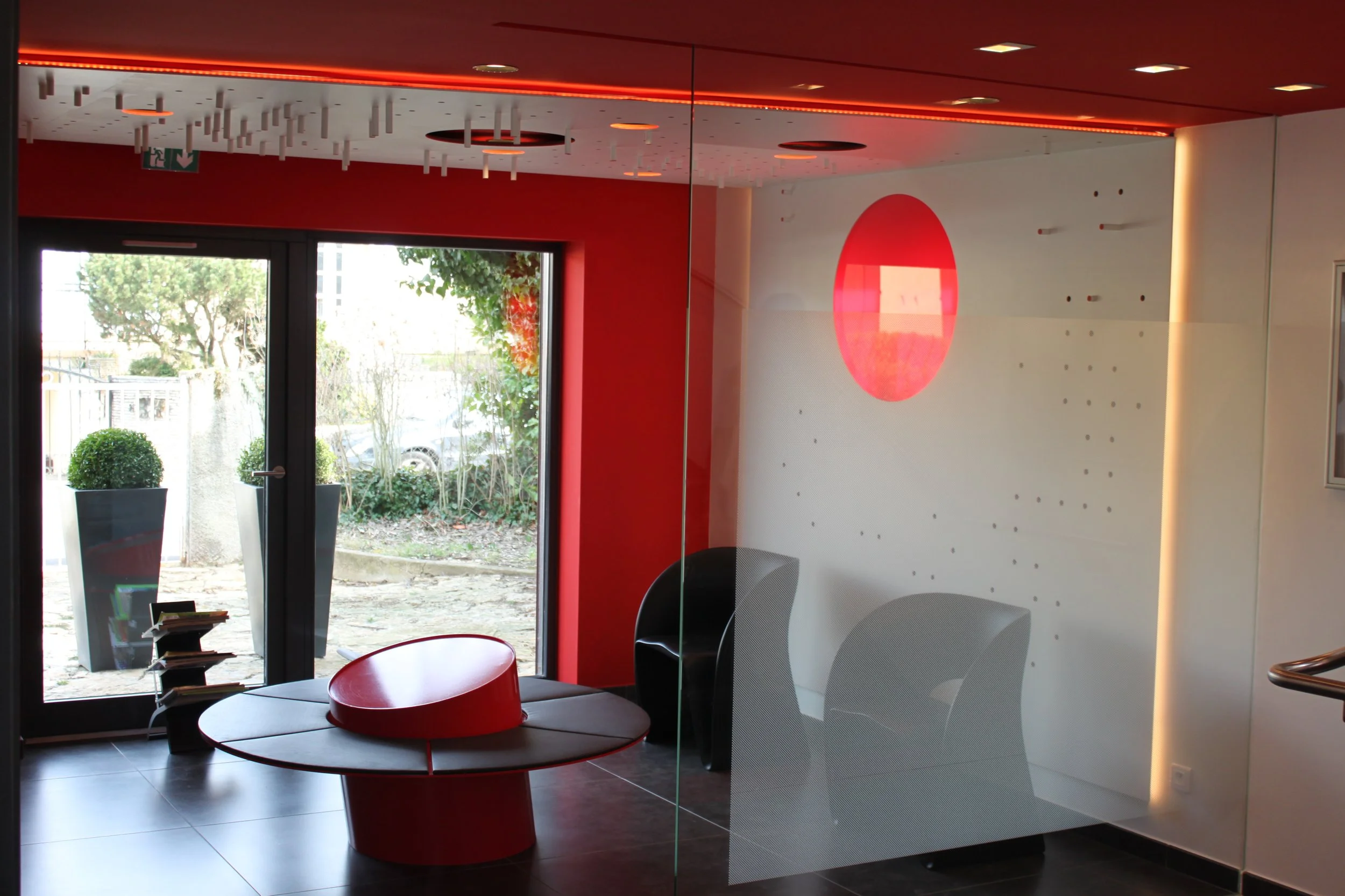 Une salle d'attente moderne avec un mobilier noir et rouge, une grande fenêtre laissant entrer la lumière naturelle, et un mur blanc avec un cercle rouge