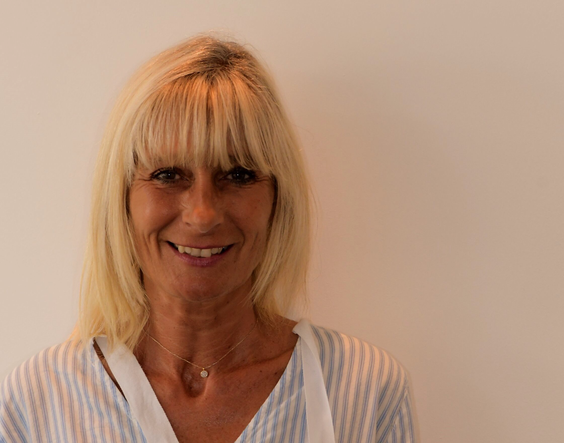 Femme blonde souriante portant une robe rayée blanc et bleu clair, avec un collier délicat, contre un fond neutre.