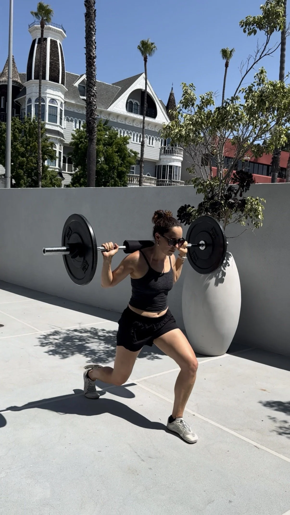 A woman wearing sunglasses, a black tank top, and black shorts is doing a lunging exercise while carrying a barbell on her shoulders outdoors on a sunny day. She is in front of a white wall with a large plant and tall palm trees in the background.