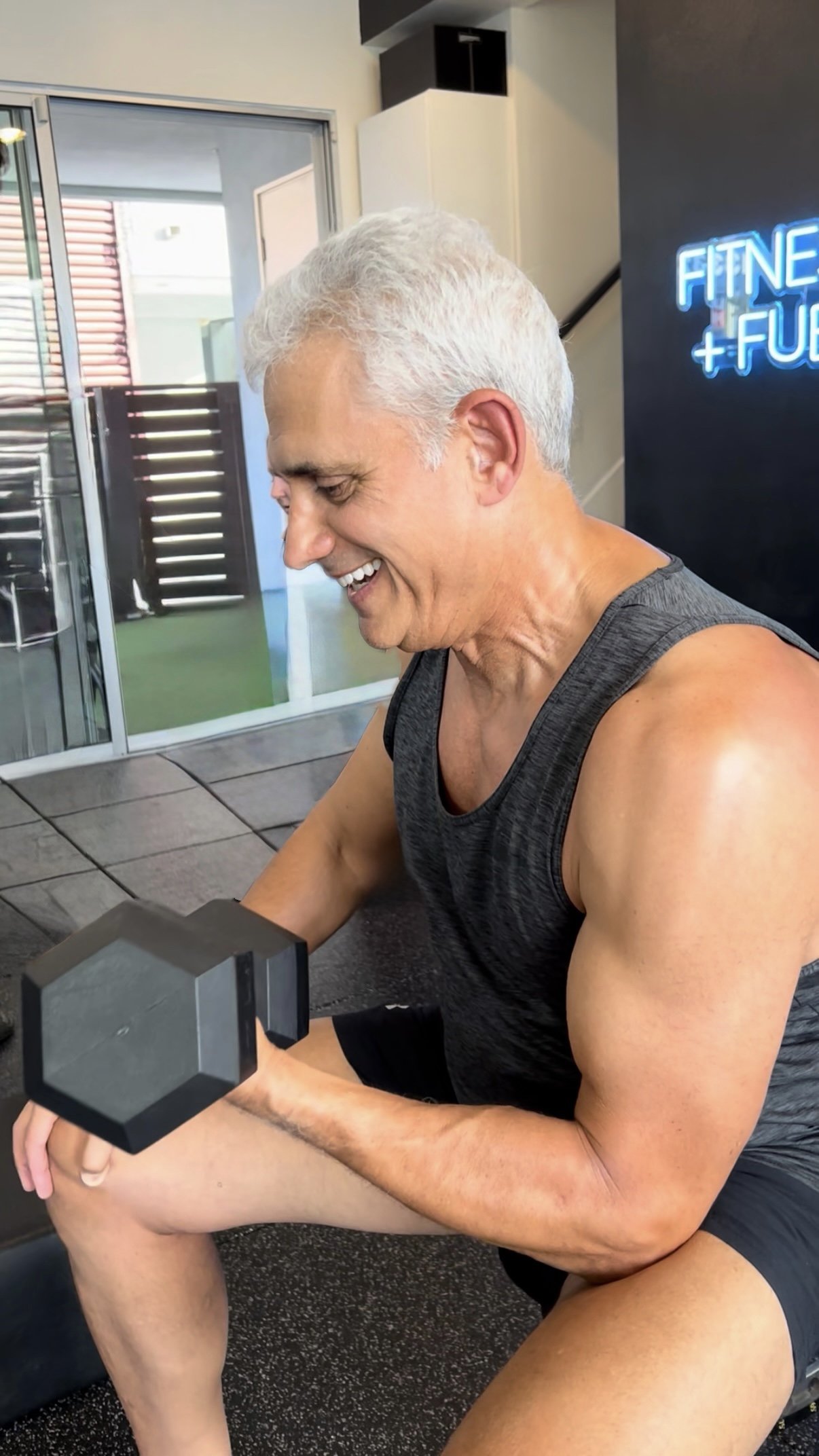 A man lying on a workout bench lifting dumbbells in a gym with fitness equipment and a mirror.