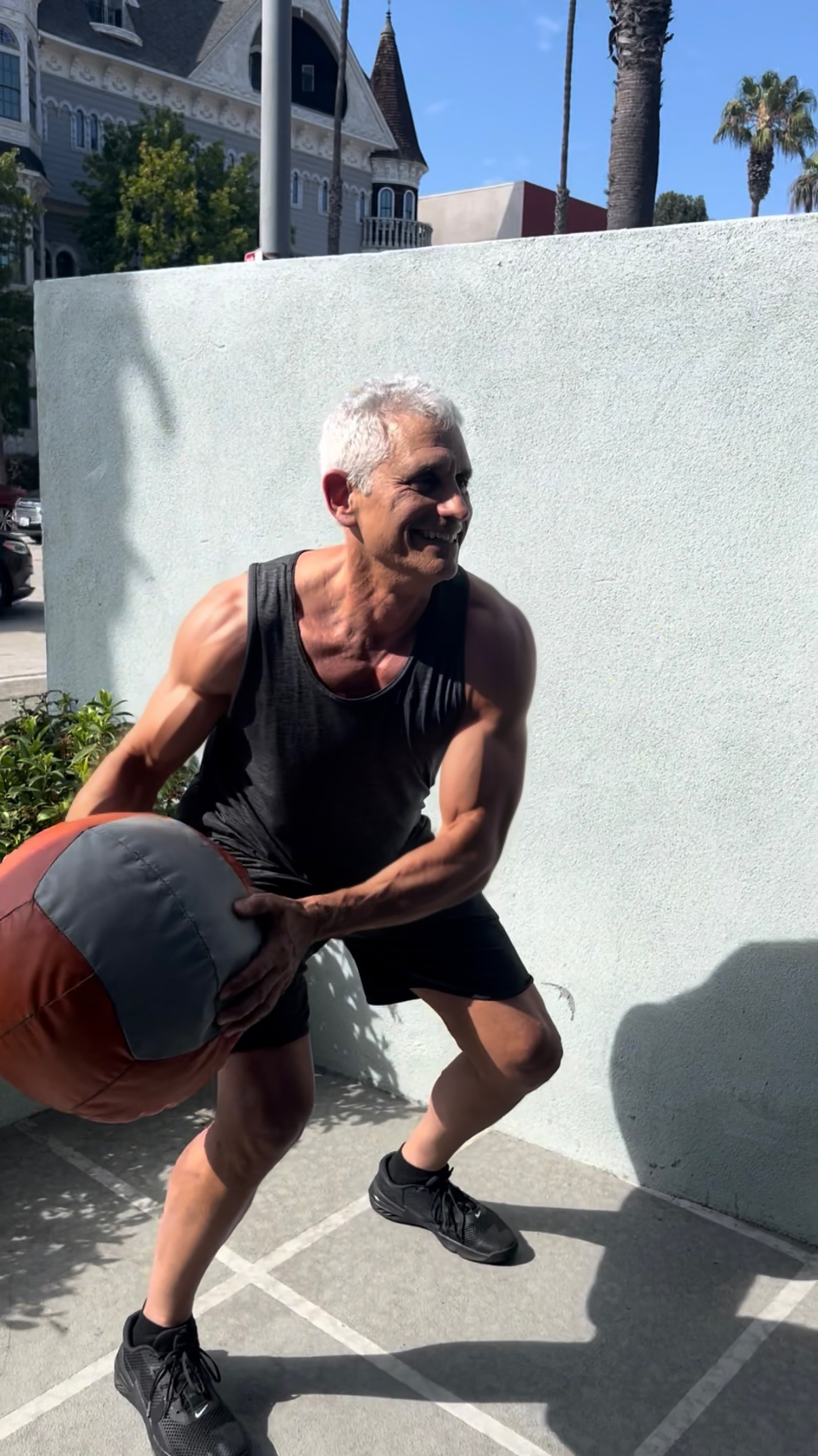 A man exercising outdoors, holding a large medicine ball with both hands, in a squat position, wearing a black tank top, black shorts, and black athletic shoes.