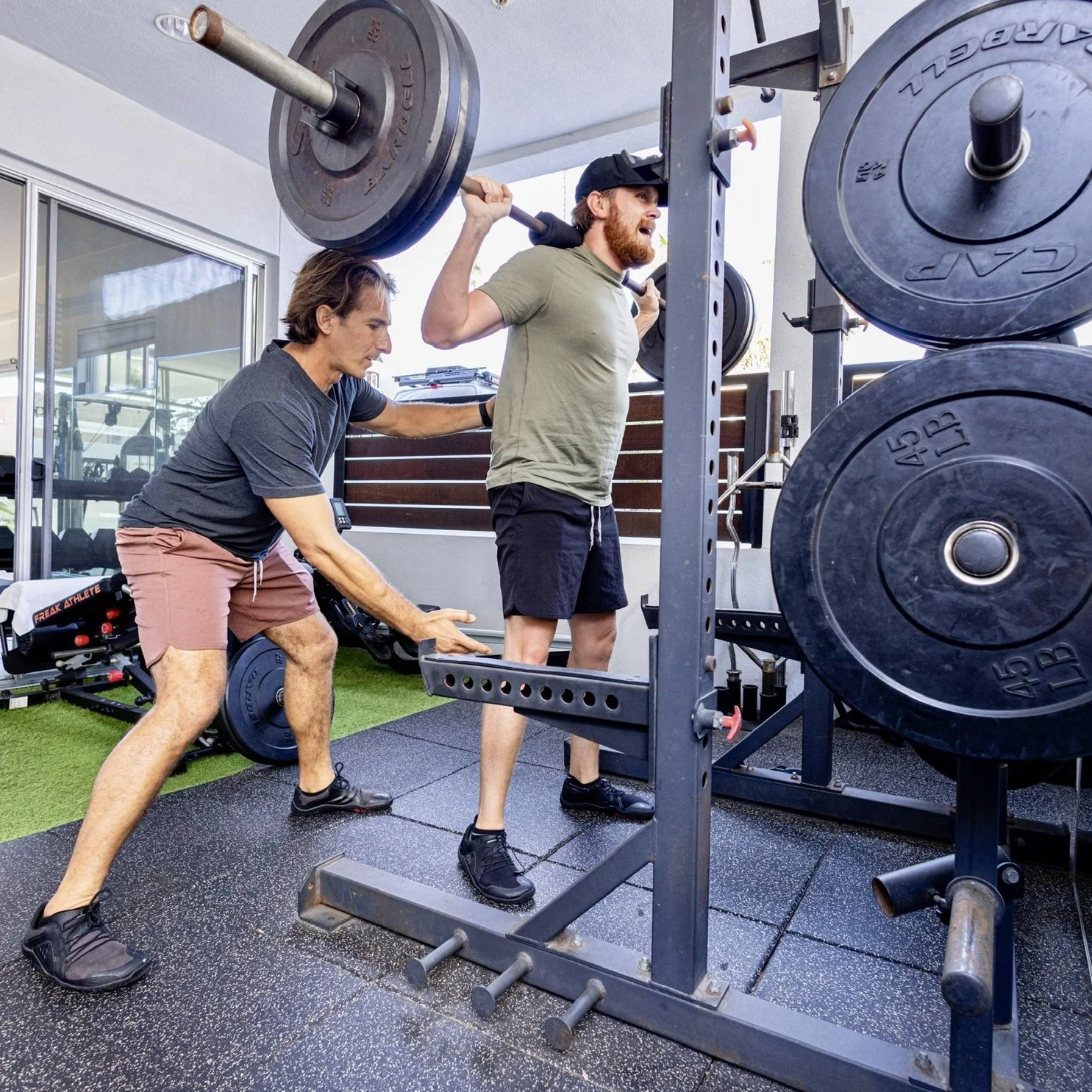 A woman doing a shoulder press exercise with a trainer assisting her, in a gym with fitness equipment and weights visible in the background.