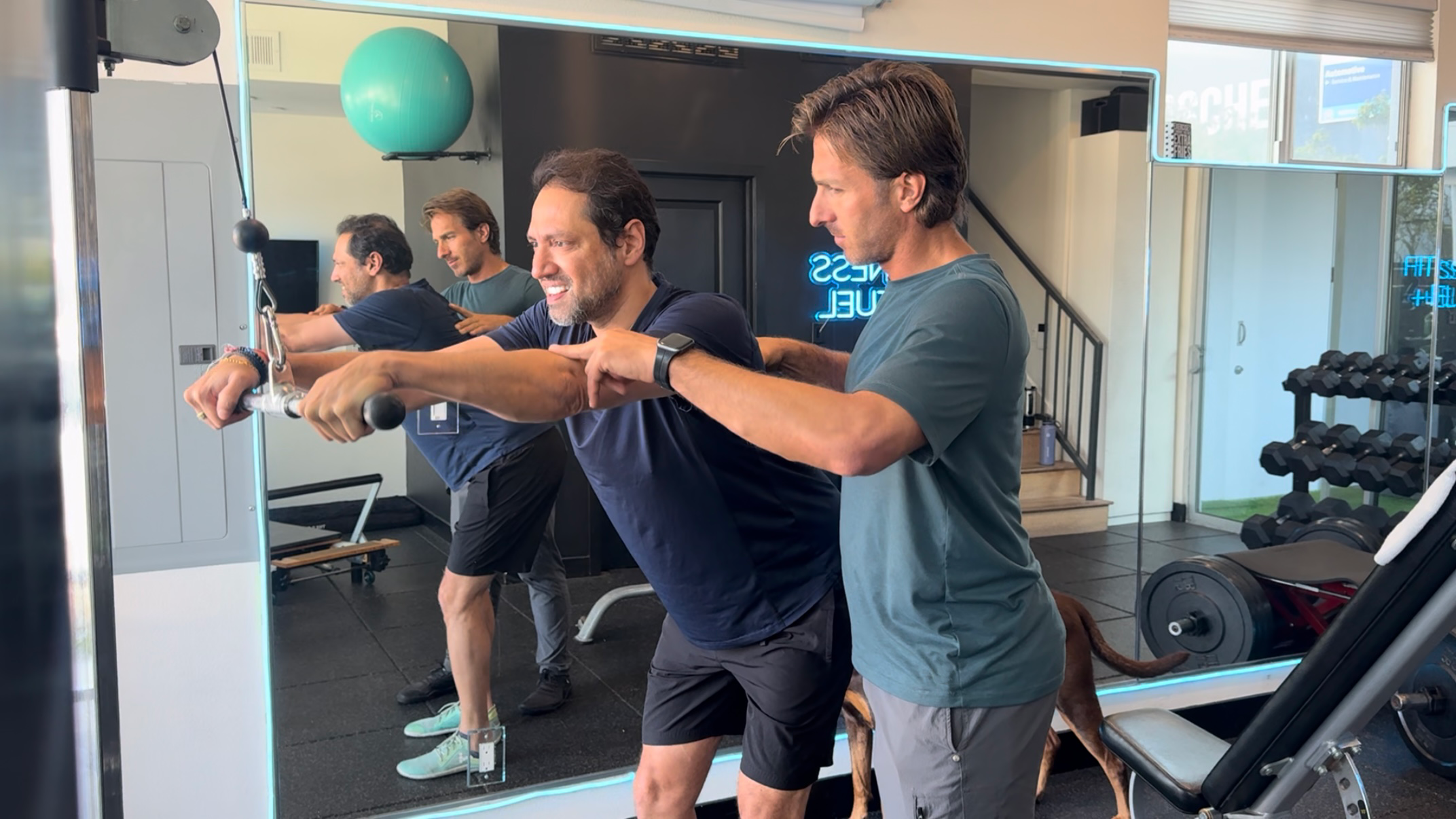 Man at Fitness and Fuel LA, stretching with the assistance of a trainer. They are in front of a mirror, and there is gym equipment like weights and a bench visible in the background.