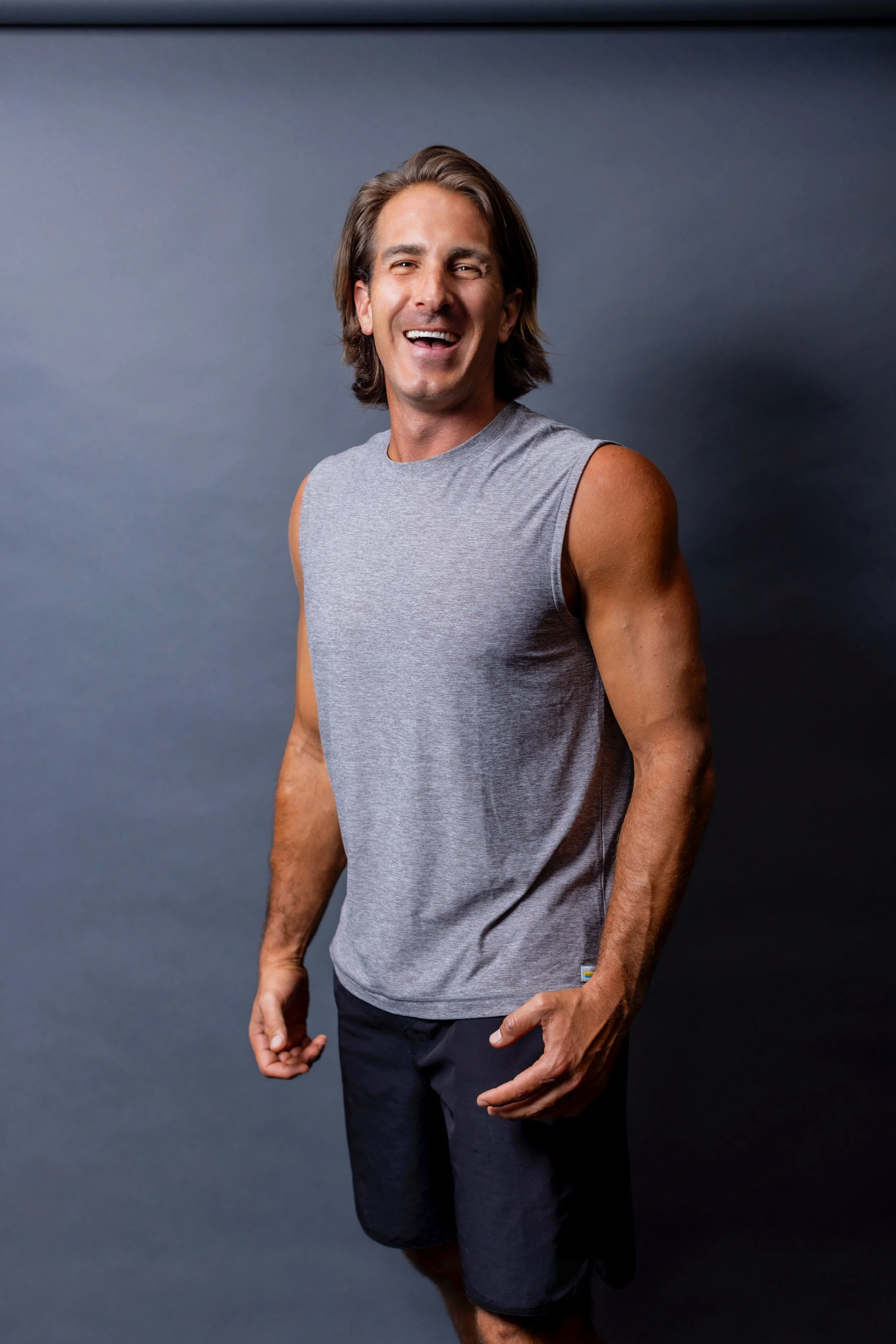 A man, Byron Paidoussi with shoulder-length brown hair, wearing a sleeveless gray athletic shirt and black shorts, smiling and standing against a plain dark background.