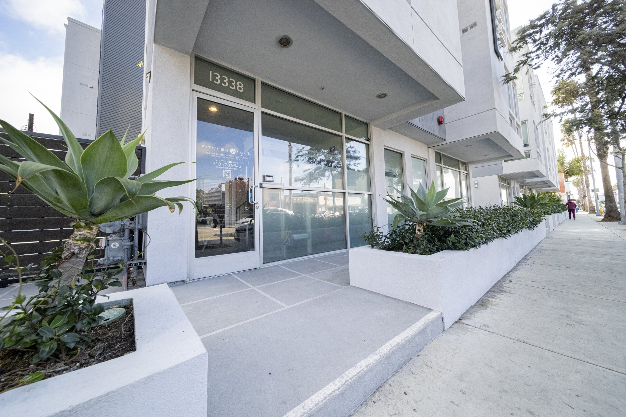 Modern building with large glass windows and doors, location number 13338, with potted agave plants and a row of smaller plants along the sidewalk.