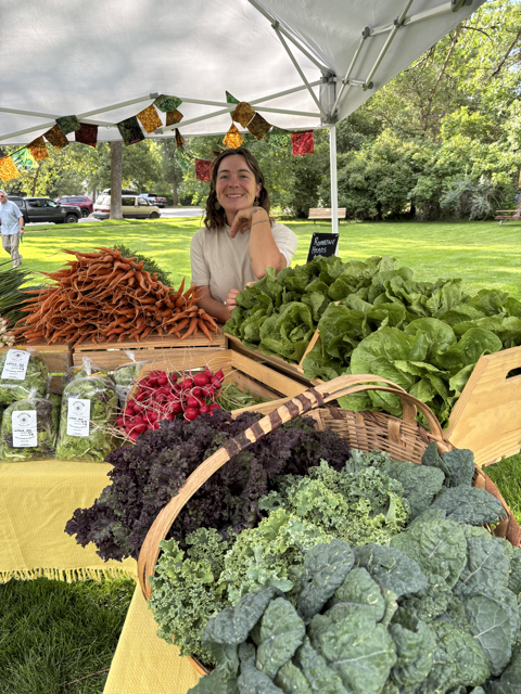 Woman at a farmers market stand with fresh vegetables including lettuce, radishes, kale, and dried peppers under a white canopy.